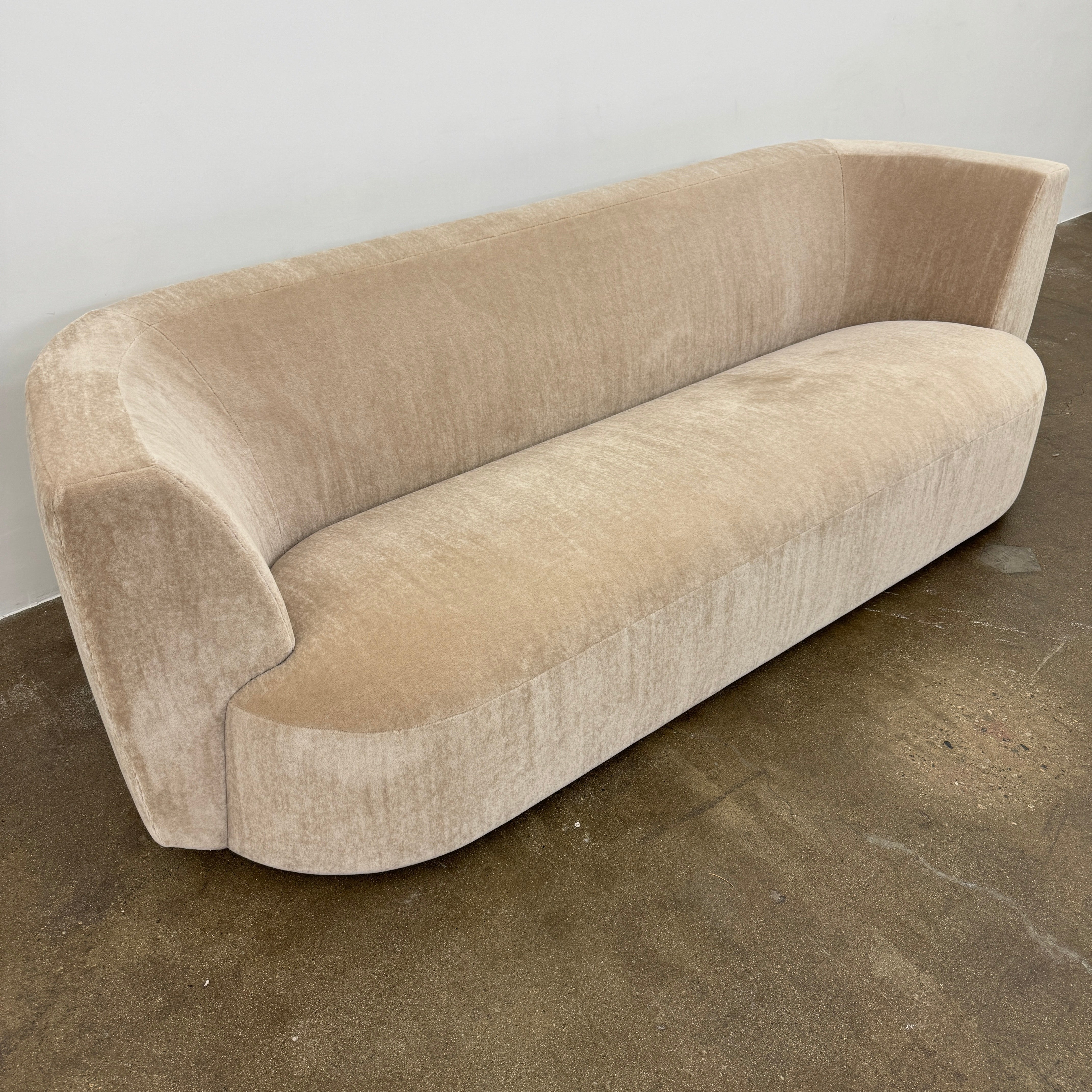 The tan, modern Holly Hunt Caste Malta Sofa with rounded edges sits stylishly on a polished concrete floor against a white wall, showcasing minimalist design inspired by Holly Hunt’s signature aesthetic.