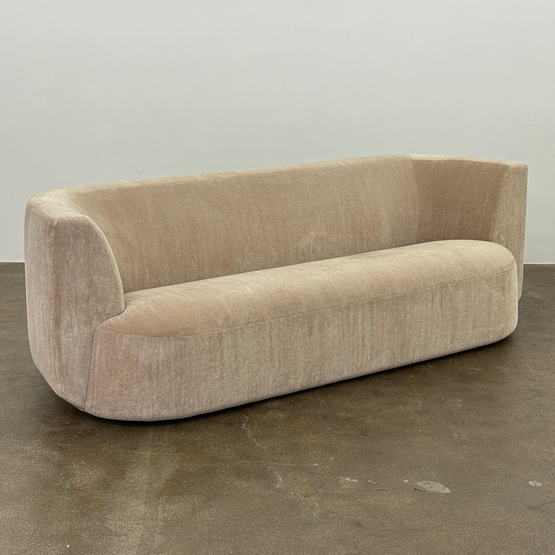 The tan, modern Holly Hunt Caste Malta Sofa with rounded edges sits stylishly on a polished concrete floor against a white wall, showcasing minimalist design inspired by Holly Hunt’s signature aesthetic.