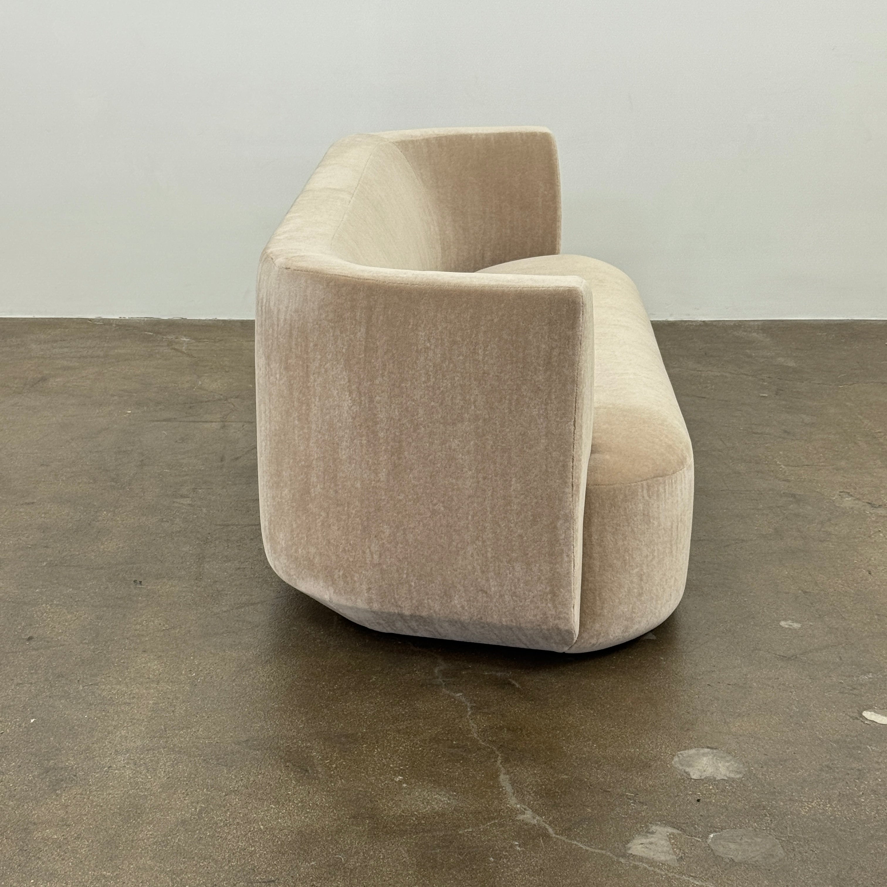 The tan, modern Holly Hunt Caste Malta Sofa with rounded edges sits stylishly on a polished concrete floor against a white wall, showcasing minimalist design inspired by Holly Hunt’s signature aesthetic.