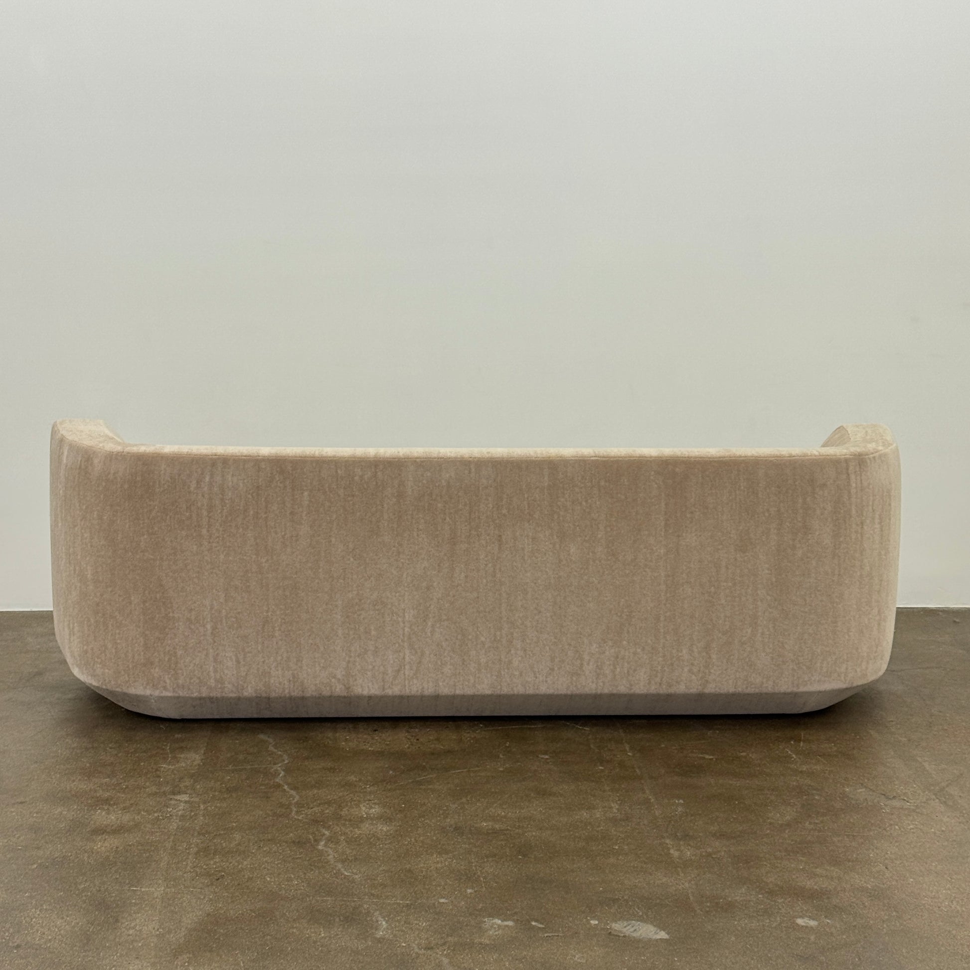 The tan, modern Holly Hunt Caste Malta Sofa with rounded edges sits stylishly on a polished concrete floor against a white wall, showcasing minimalist design inspired by Holly Hunt’s signature aesthetic.