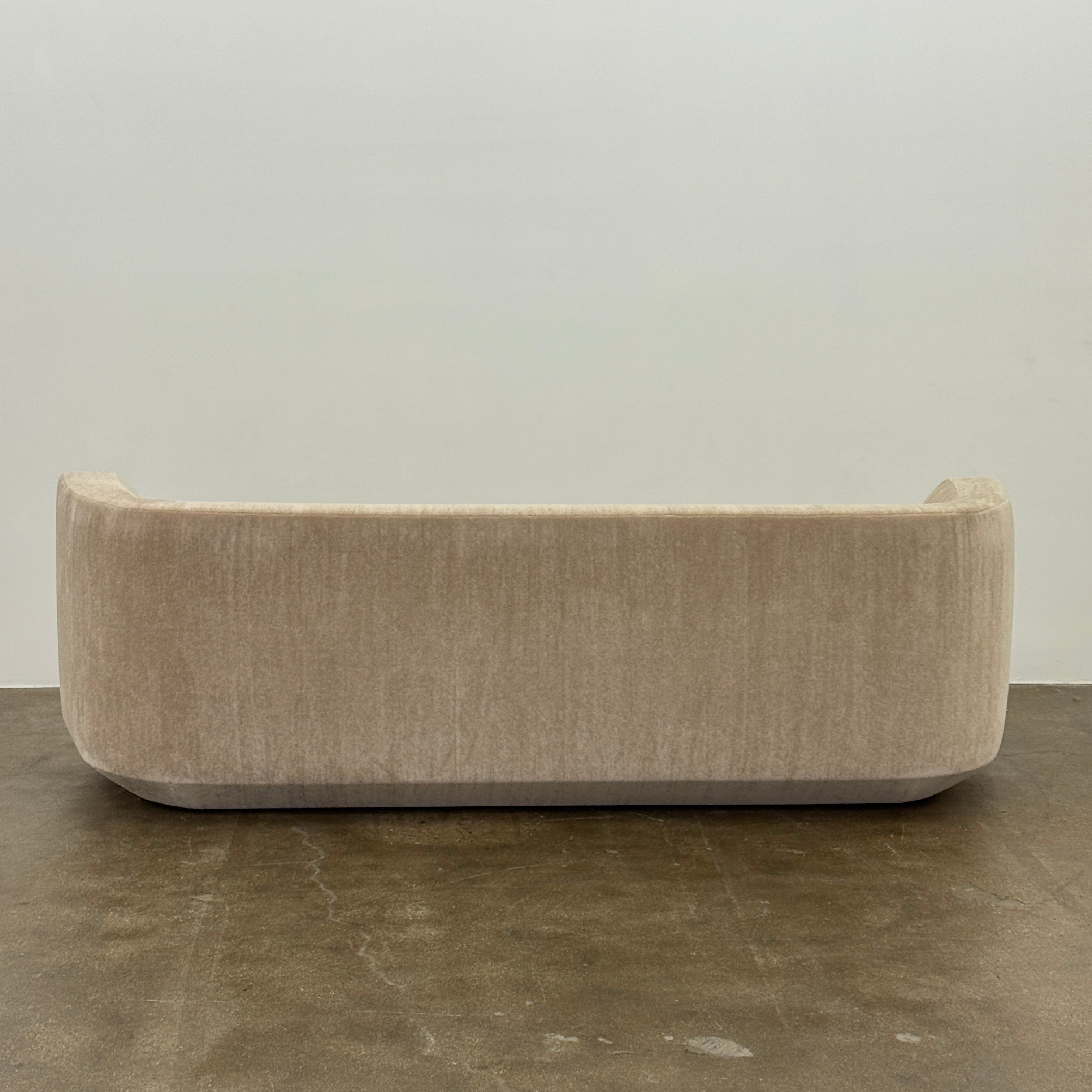 The tan, modern Holly Hunt Caste Malta Sofa with rounded edges sits stylishly on a polished concrete floor against a white wall, showcasing minimalist design inspired by Holly Hunt’s signature aesthetic.