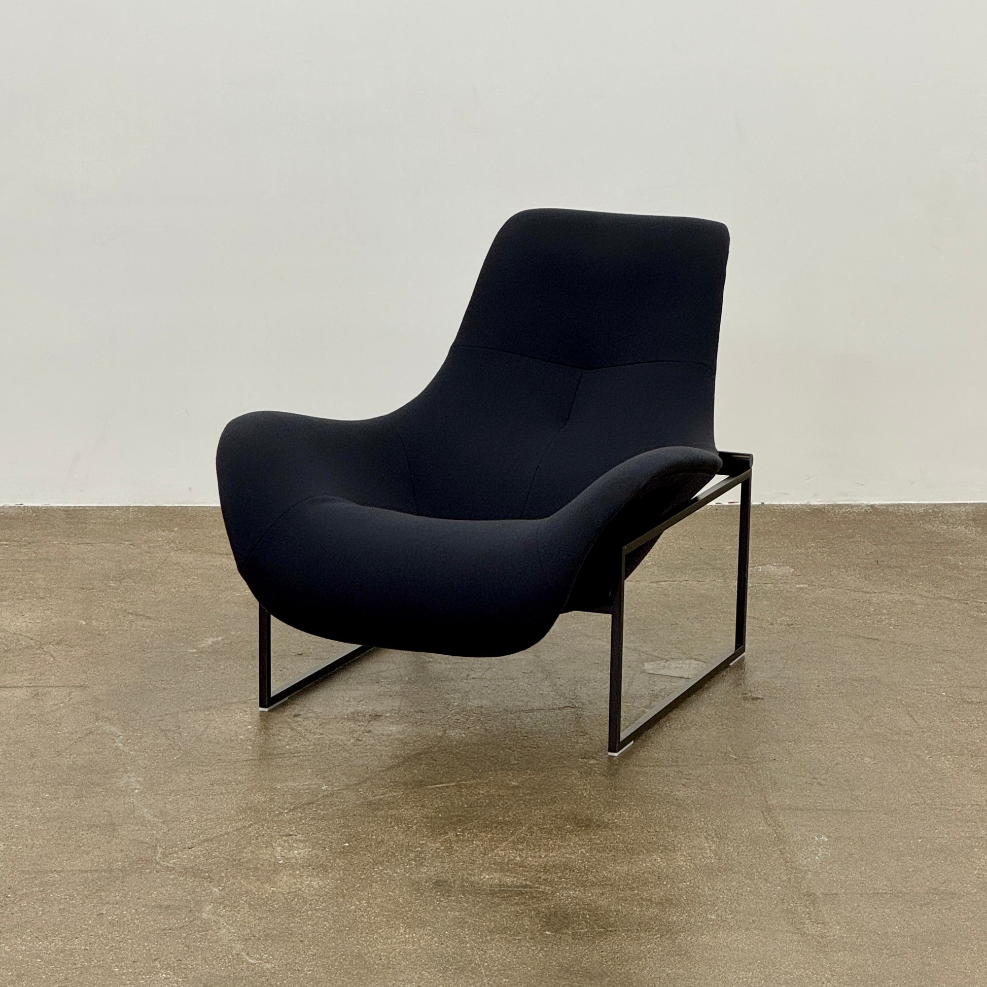A B&B Italia Mart Relax Armchair with tilting mechanism and fabric seat by B&B Italia, featuring curved cushioned seating and a minimalist metal frame, sits on a concrete floor against a plain light-colored wall.