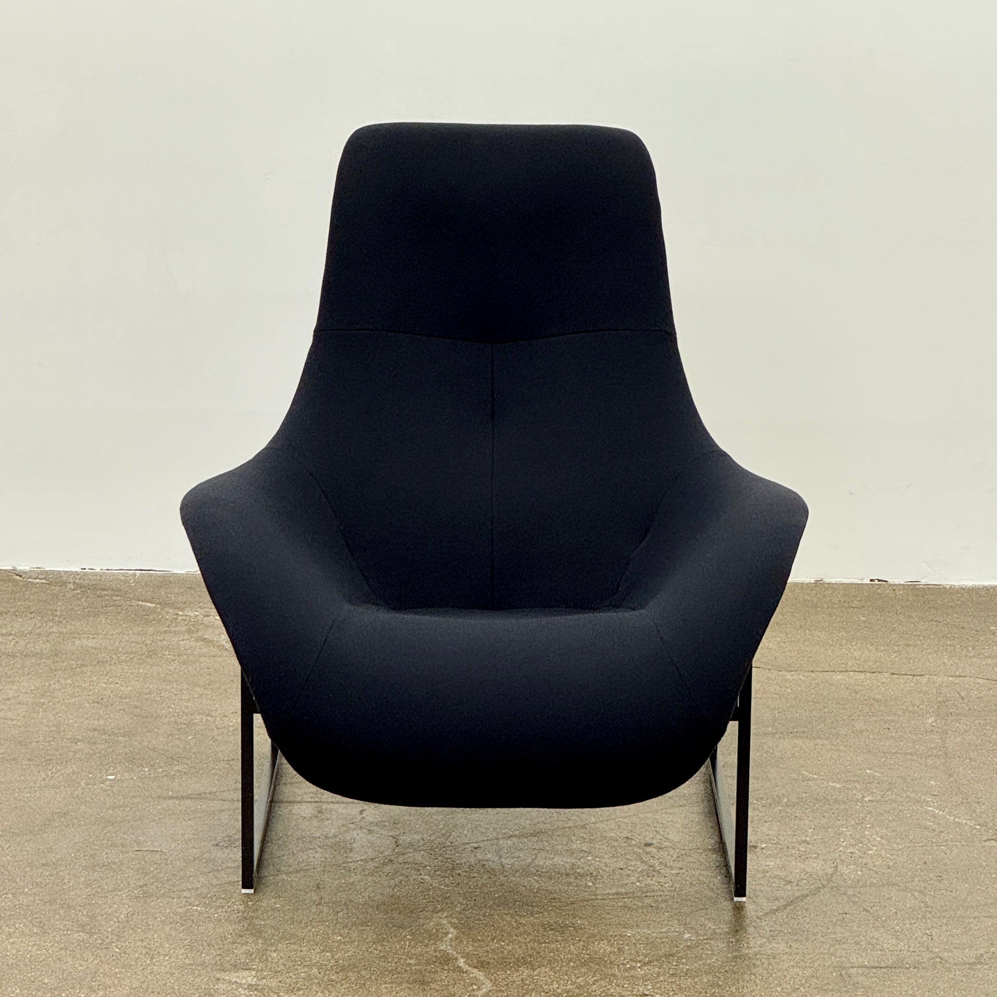 A B&B Italia Mart Relax Armchair with tilting mechanism and fabric seat by B&B Italia, featuring curved cushioned seating and a minimalist metal frame, sits on a concrete floor against a plain light-colored wall.