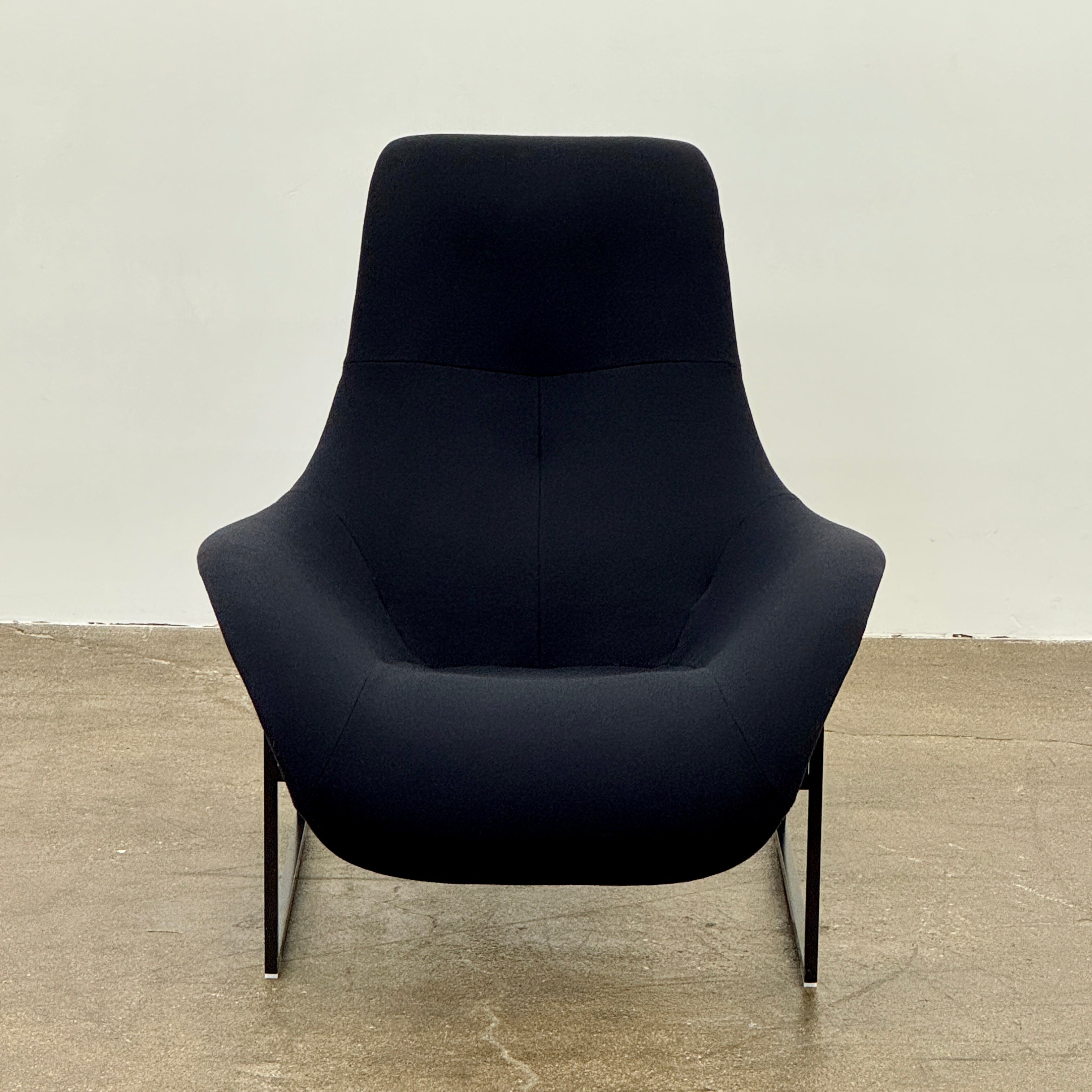 A B&B Italia Mart Relax Armchair with tilting mechanism and fabric seat by B&B Italia, featuring curved cushioned seating and a minimalist metal frame, sits on a concrete floor against a plain light-colored wall.