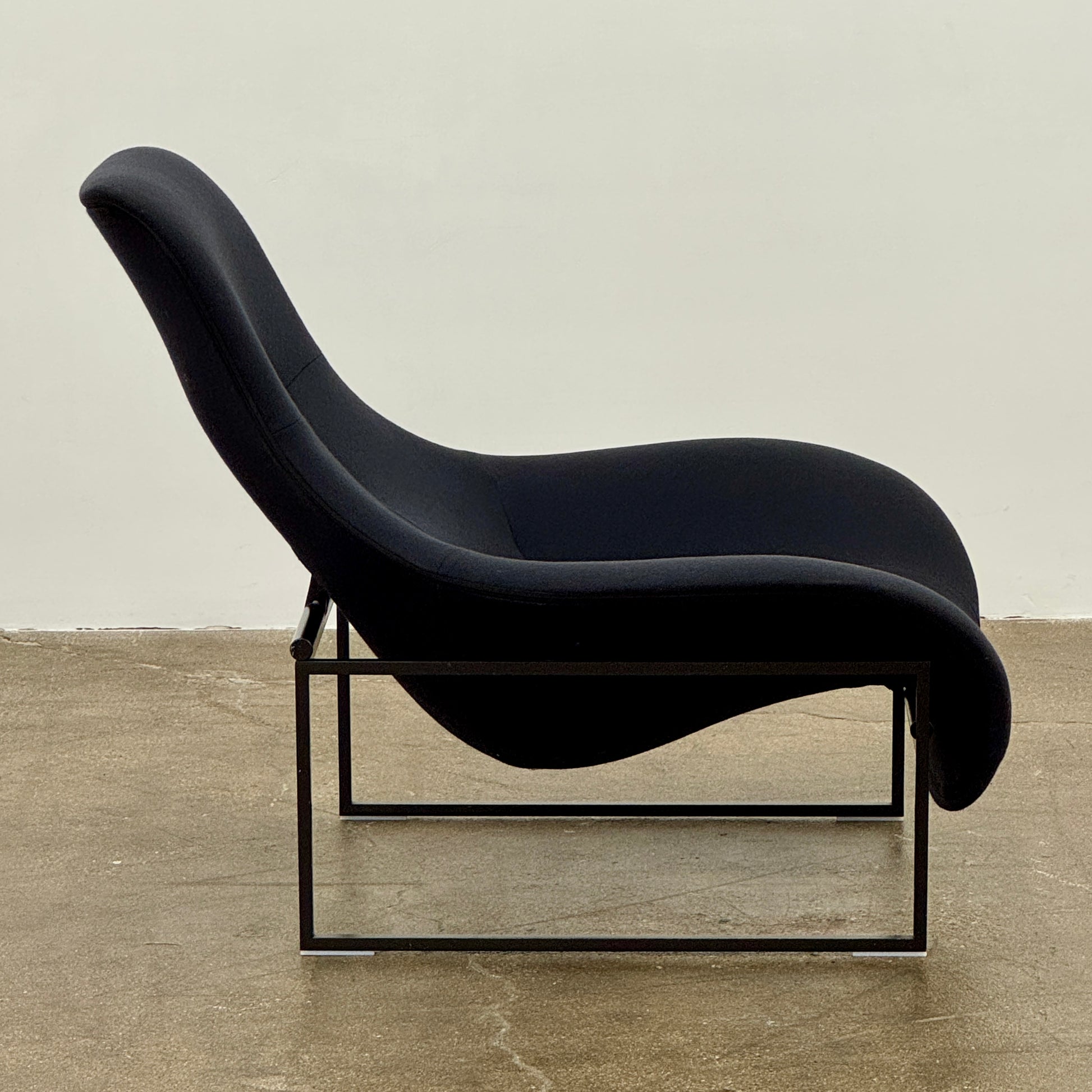 A B&B Italia Mart Relax Armchair with tilting mechanism and fabric seat by B&B Italia, featuring curved cushioned seating and a minimalist metal frame, sits on a concrete floor against a plain light-colored wall.