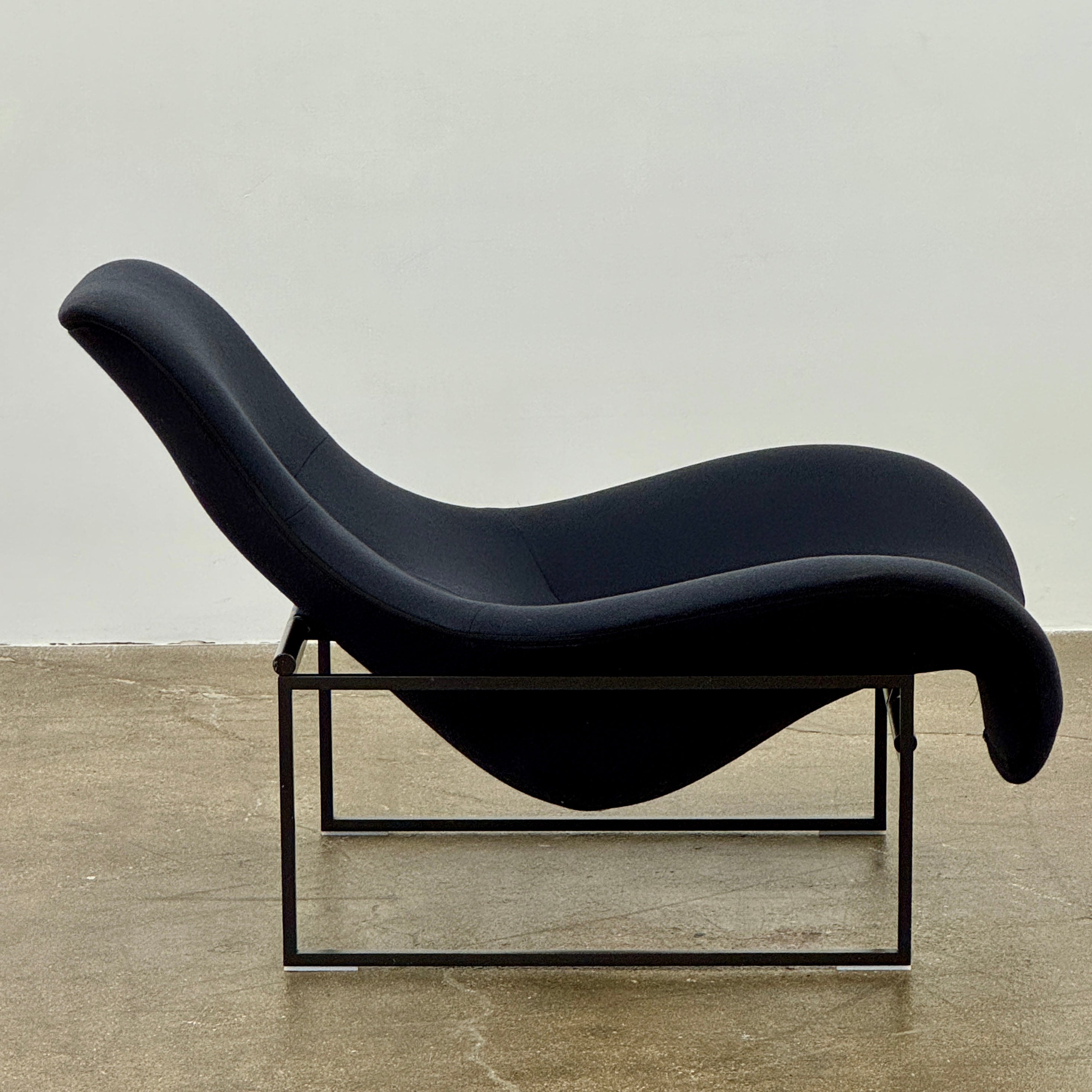 A B&B Italia Mart Relax Armchair with tilting mechanism and fabric seat by B&B Italia, featuring curved cushioned seating and a minimalist metal frame, sits on a concrete floor against a plain light-colored wall.