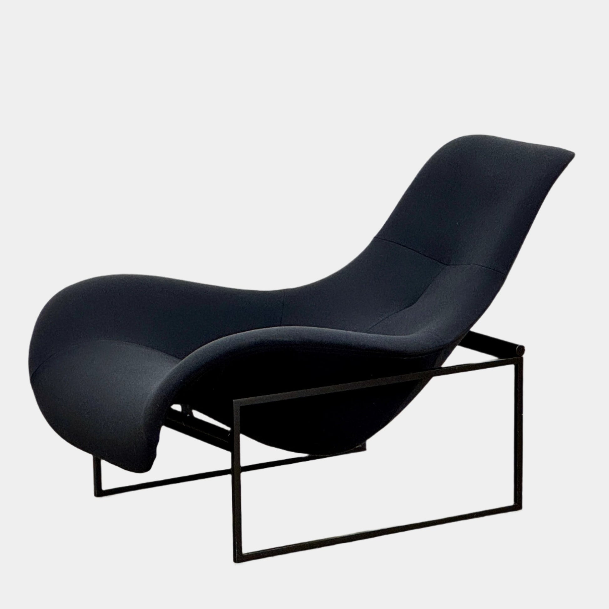 A B&B Italia Mart Relax Armchair with tilting mechanism and fabric seat by B&B Italia, featuring curved cushioned seating and a minimalist metal frame, sits on a concrete floor against a plain light-colored wall.