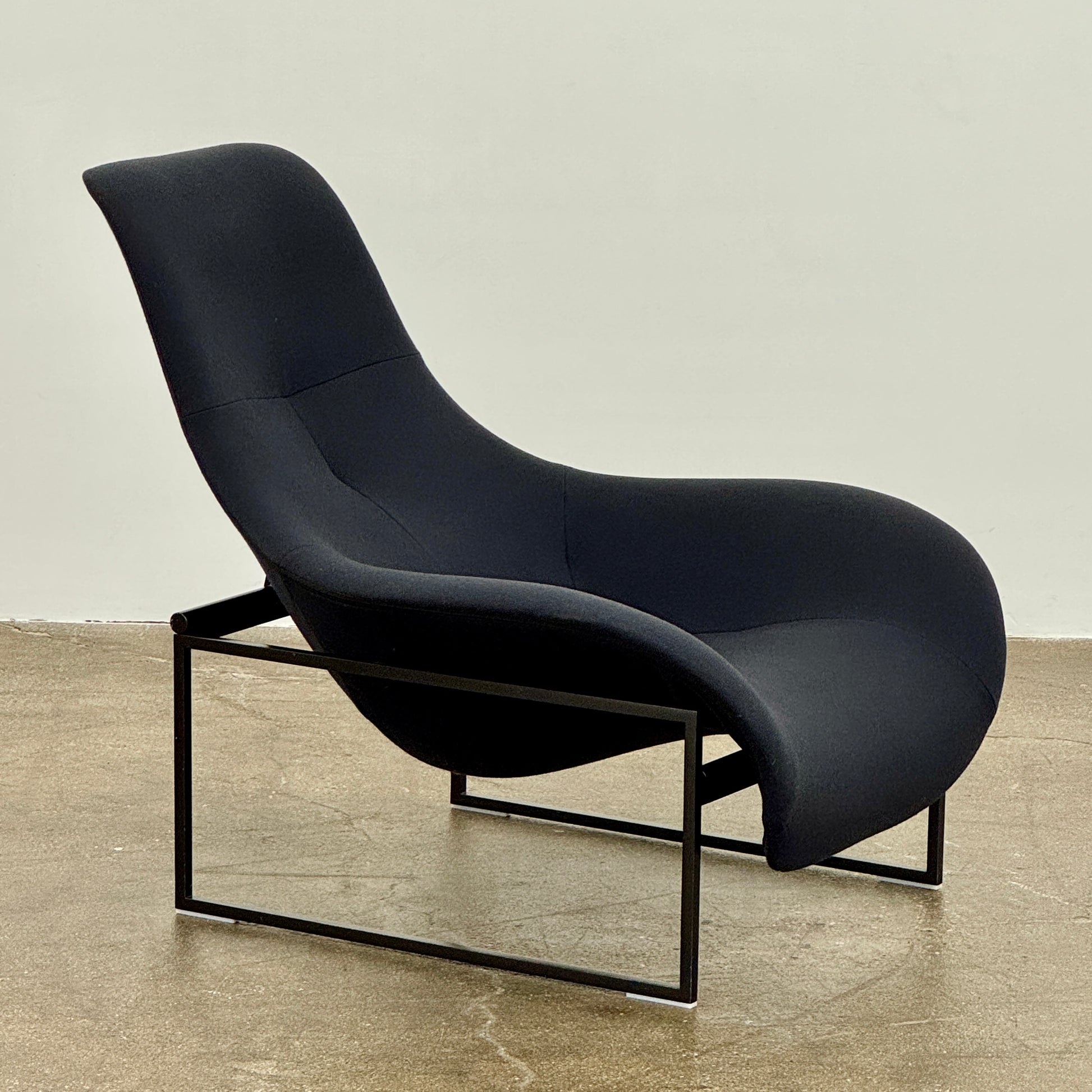 A B&B Italia Mart Relax Armchair with tilting mechanism and fabric seat by B&B Italia, featuring curved cushioned seating and a minimalist metal frame, sits on a concrete floor against a plain light-colored wall.