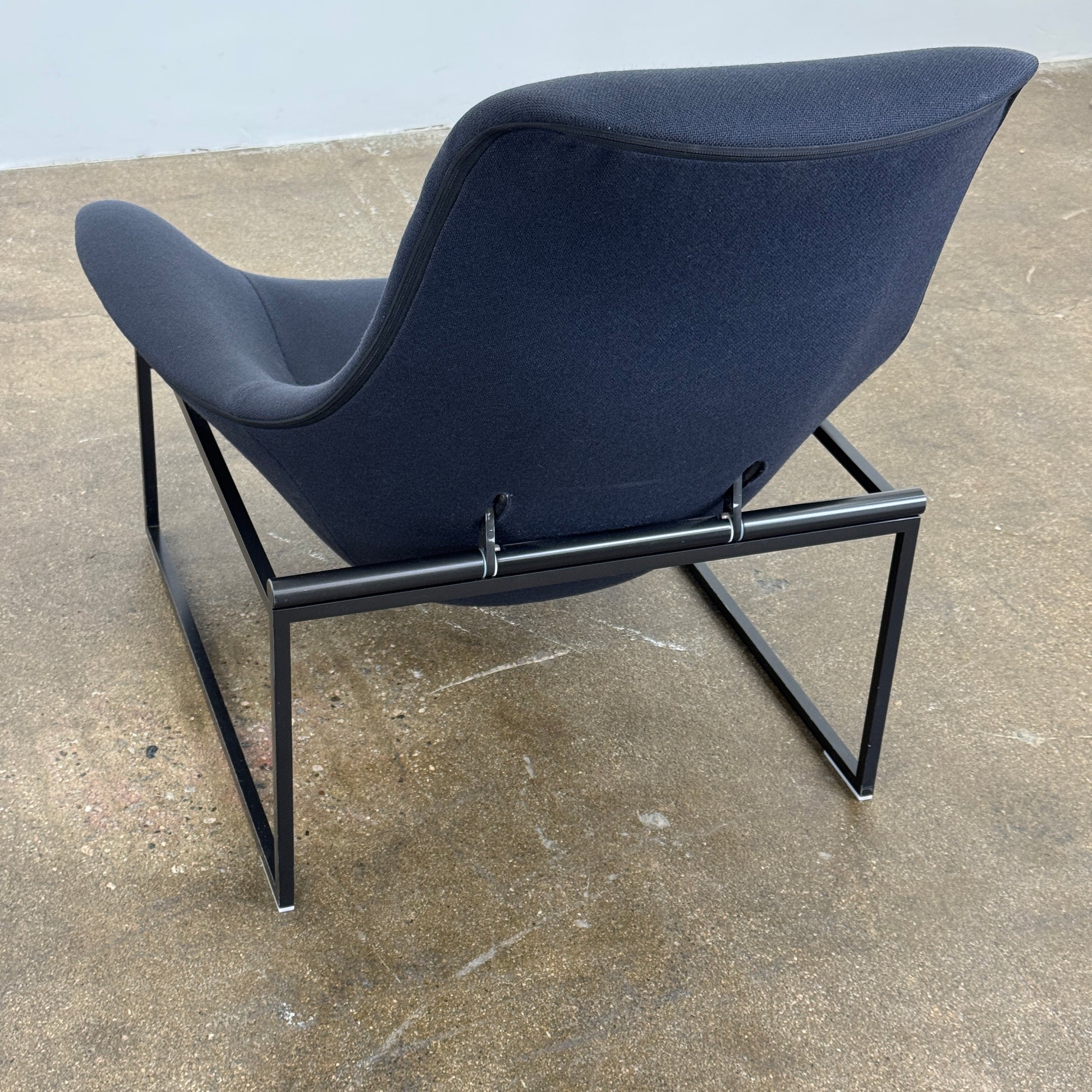 A B&B Italia Mart Relax Armchair with tilting mechanism and fabric seat by B&B Italia, featuring curved cushioned seating and a minimalist metal frame, sits on a concrete floor against a plain light-colored wall.