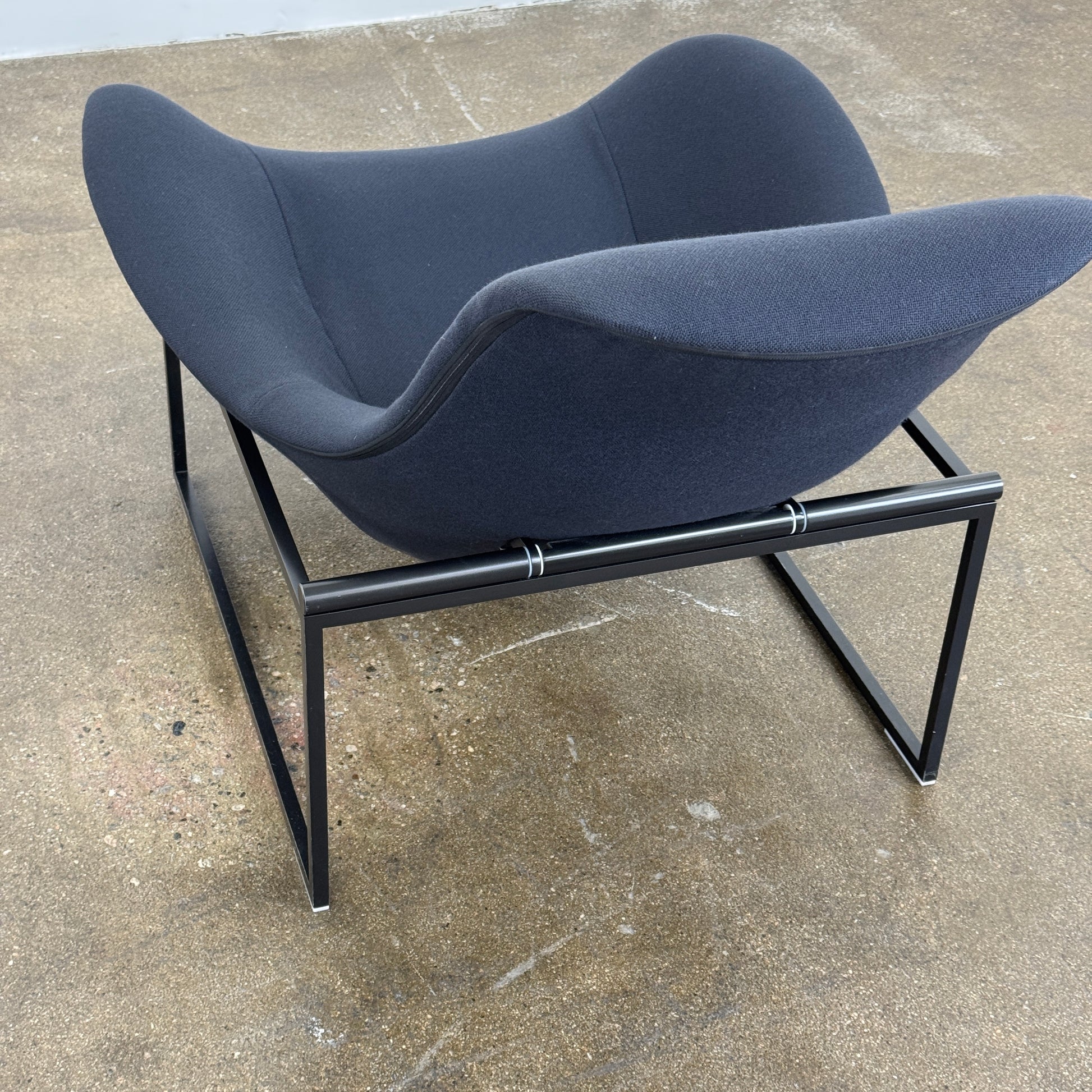 A B&B Italia Mart Relax Armchair with tilting mechanism and fabric seat by B&B Italia, featuring curved cushioned seating and a minimalist metal frame, sits on a concrete floor against a plain light-colored wall.