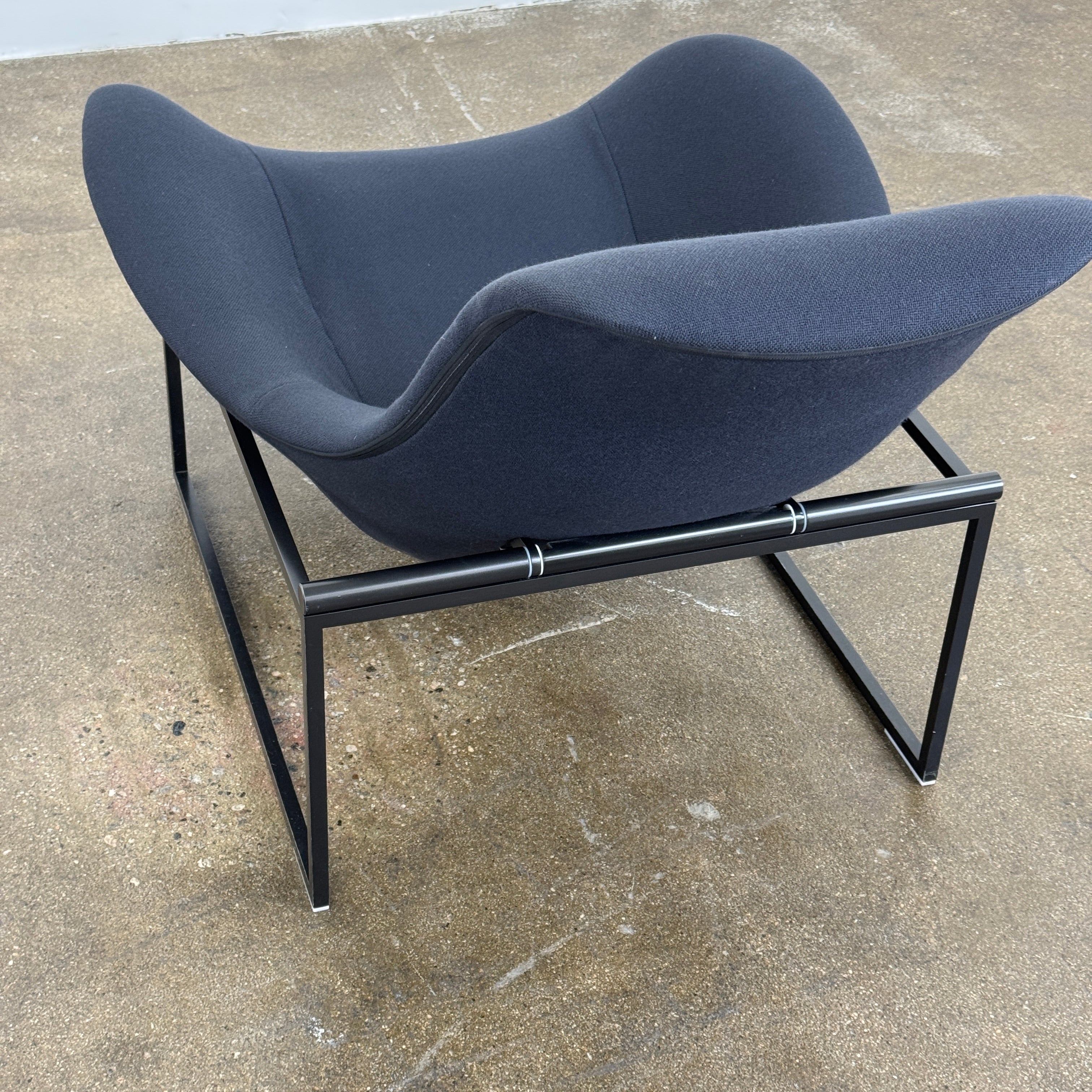 A B&B Italia Mart Relax Armchair with tilting mechanism and fabric seat by B&B Italia, featuring curved cushioned seating and a minimalist metal frame, sits on a concrete floor against a plain light-colored wall.