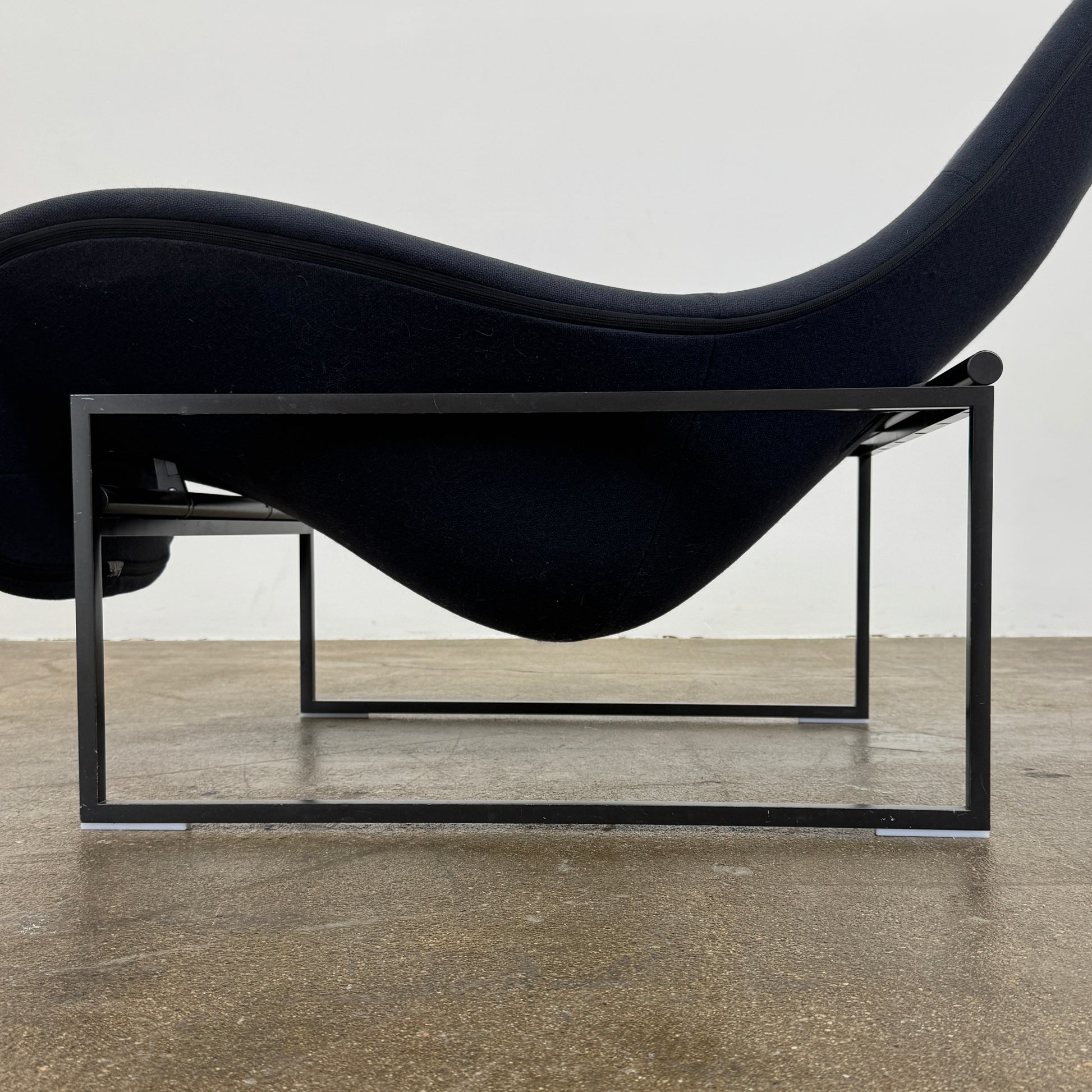 A B&B Italia Mart Relax Armchair with tilting mechanism and fabric seat by B&B Italia, featuring curved cushioned seating and a minimalist metal frame, sits on a concrete floor against a plain light-colored wall.