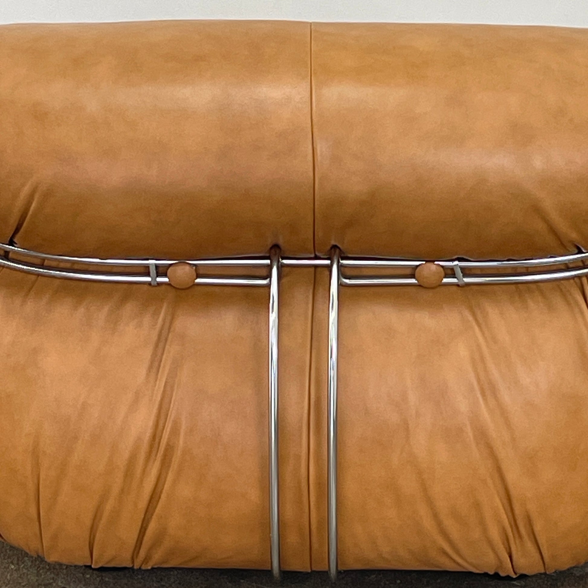 A set of two tan leather lounge chairs, echoing the vintage 1970s Cassina Soriana designs by Afra & Tobia Scarpa, showcases tufted backrests and tubular metal frames on a plain white background.