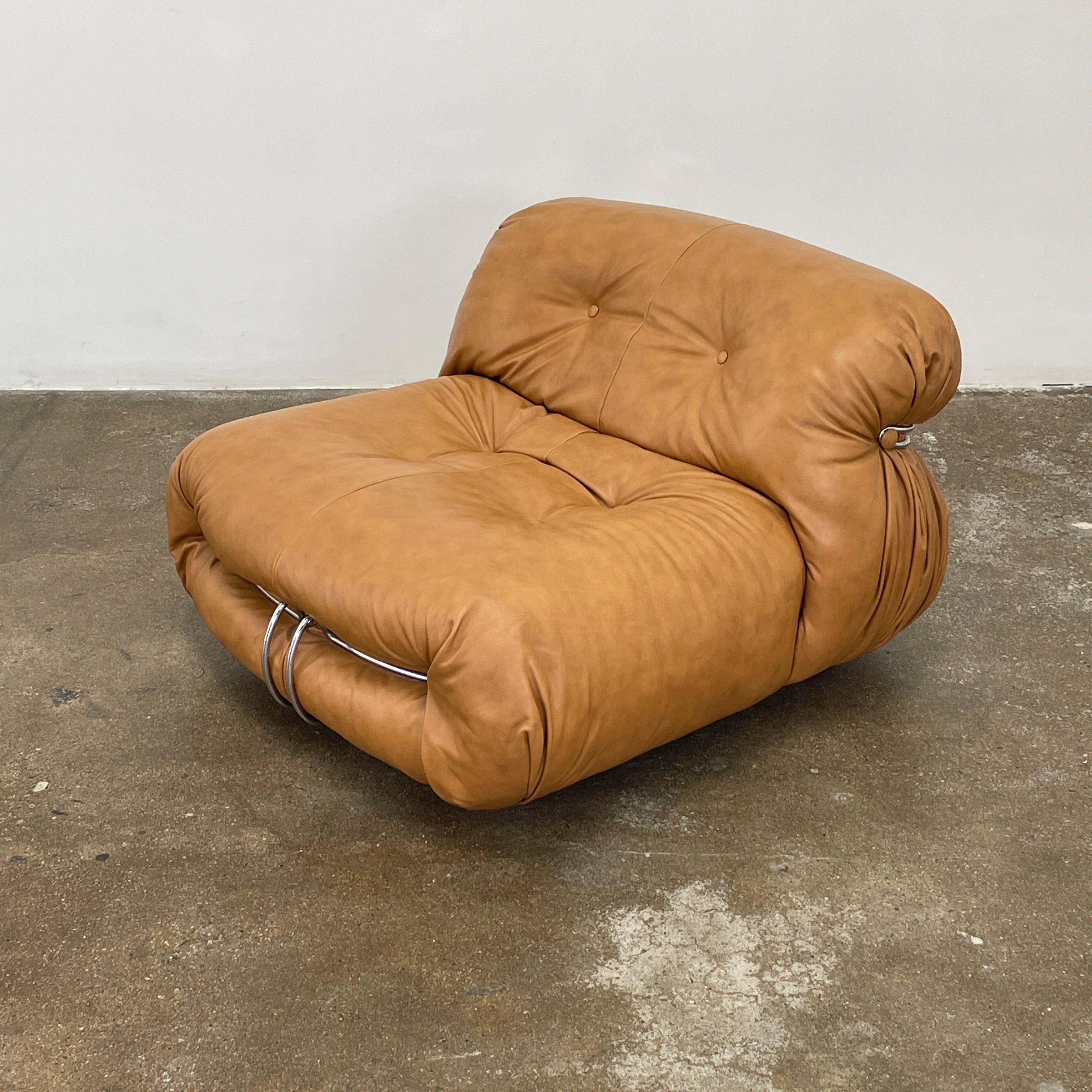 A set of two tan leather lounge chairs, echoing the vintage 1970s Cassina Soriana designs by Afra & Tobia Scarpa, showcases tufted backrests and tubular metal frames on a plain white background.
