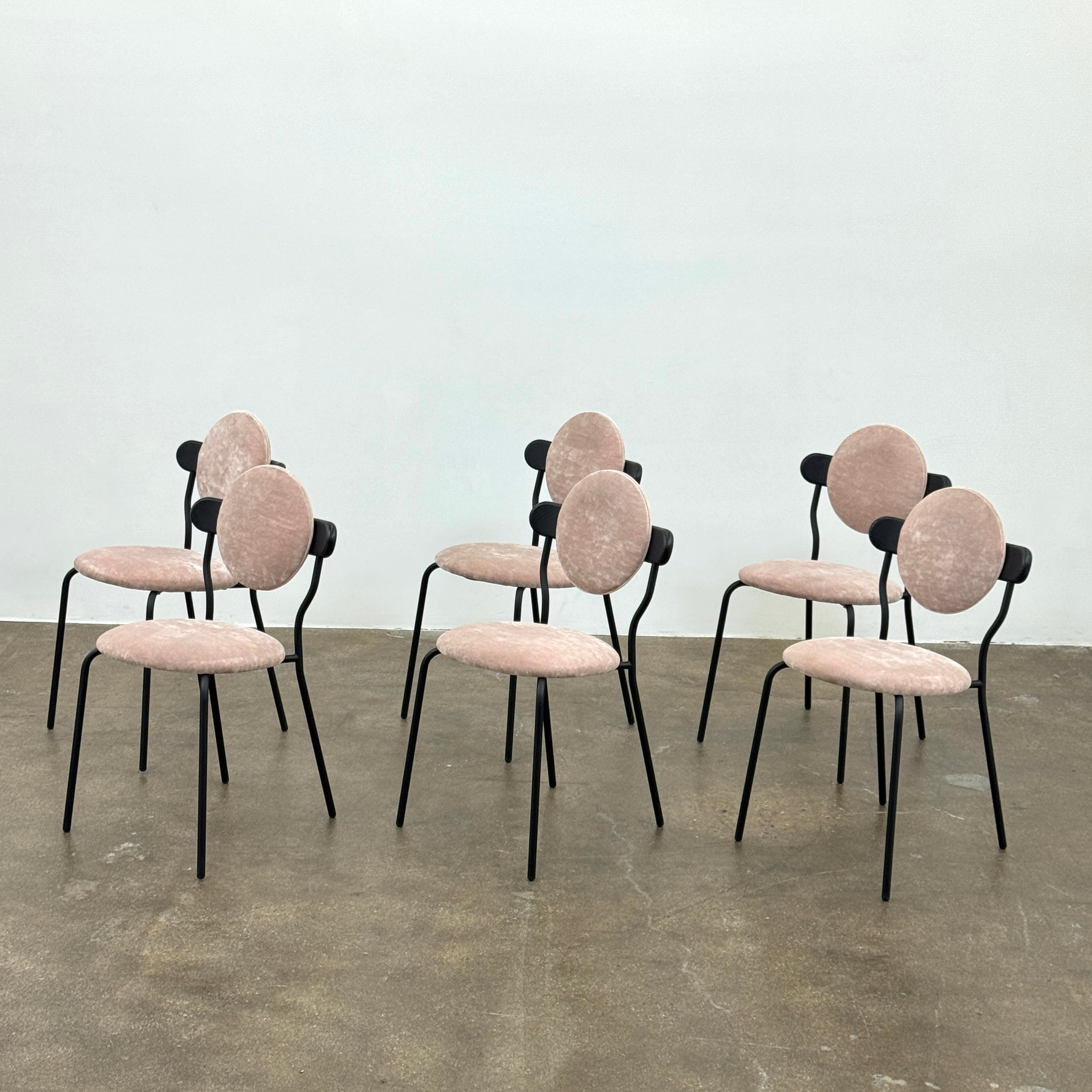 Six Modern Resale La Chance Planet Dining Chairs, set of 6, feature round light pink velvet seats and black metal legs, arranged in two rows on a bare concrete floor against a white wall—perfect for adding modern elegance.