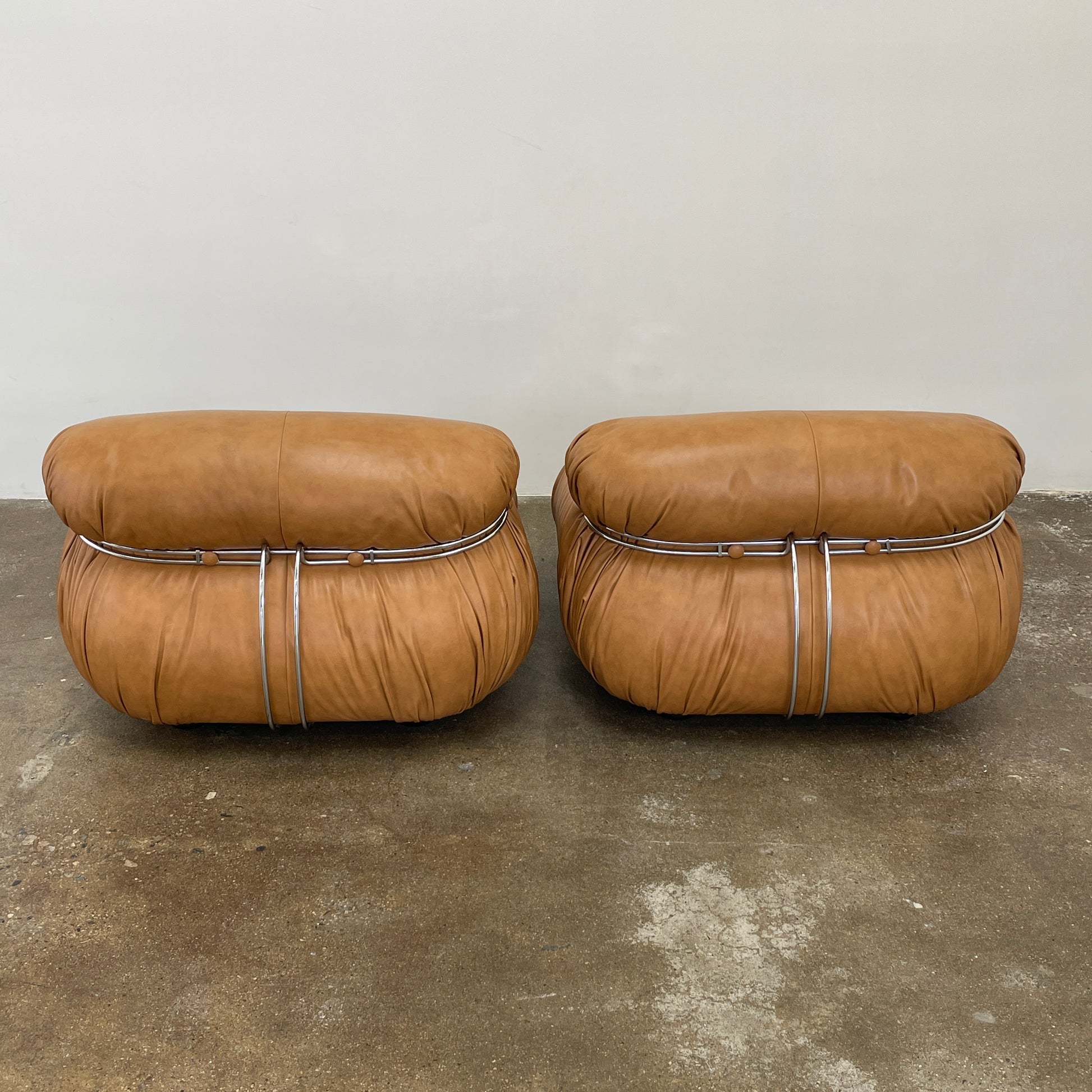 A set of two tan leather lounge chairs, echoing the vintage 1970s Cassina Soriana designs by Afra & Tobia Scarpa, showcases tufted backrests and tubular metal frames on a plain white background.