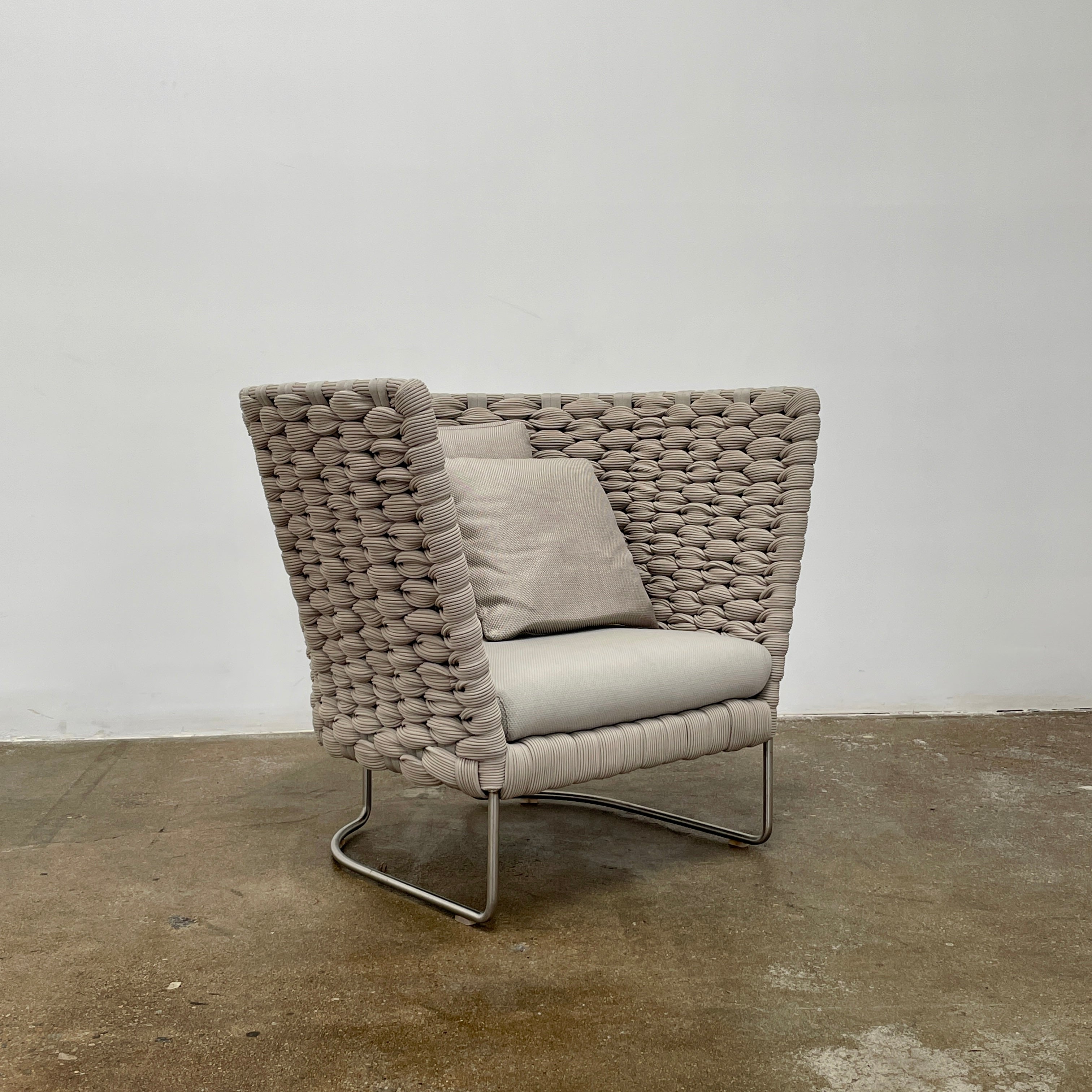 The Paola Lenti Ami Outdoor Armchair by Paola Lenti features a large weave frame, light gray cushions, and a metal base, all set against a plain white background.