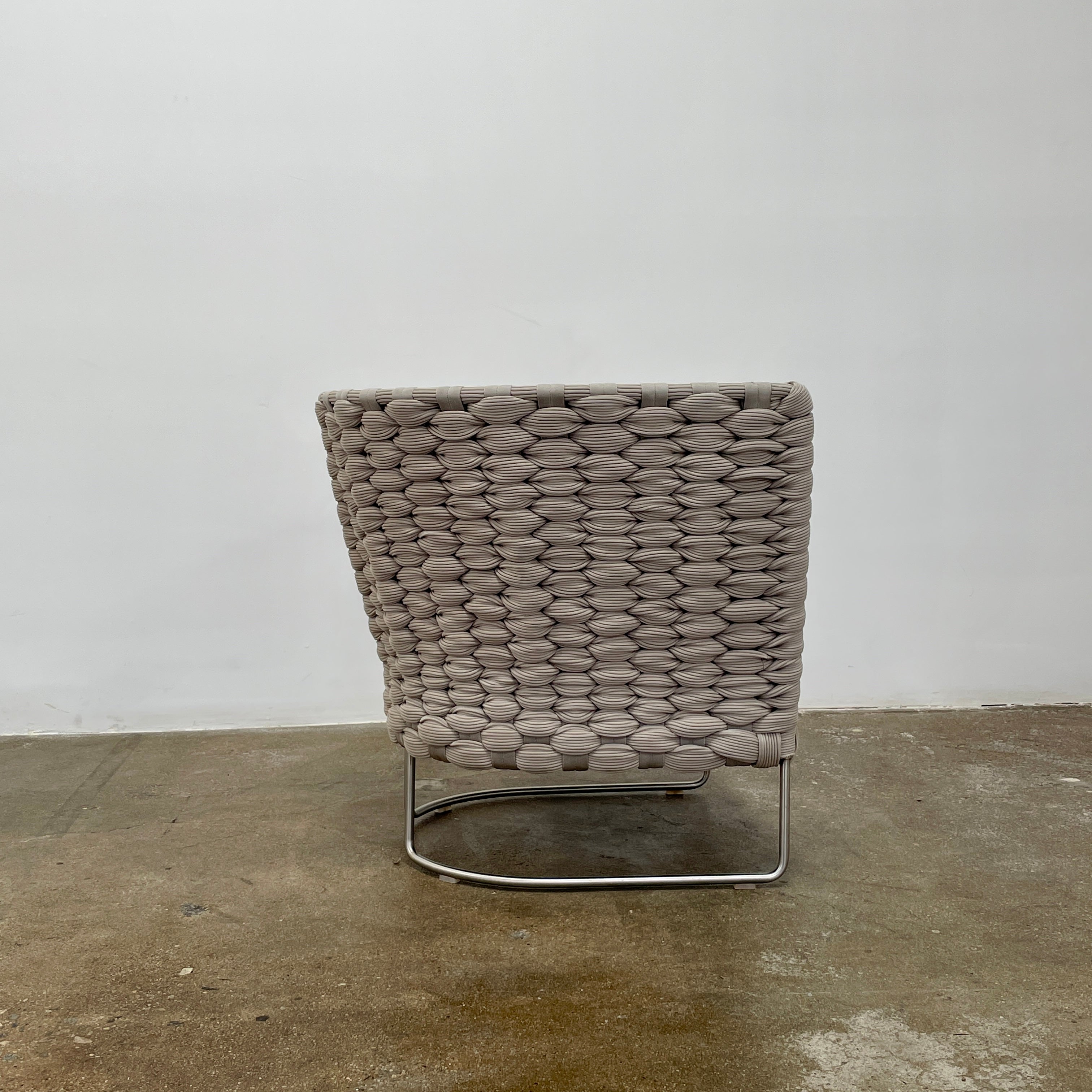 The Paola Lenti Ami Outdoor Armchair by Paola Lenti features a large weave frame, light gray cushions, and a metal base, all set against a plain white background.