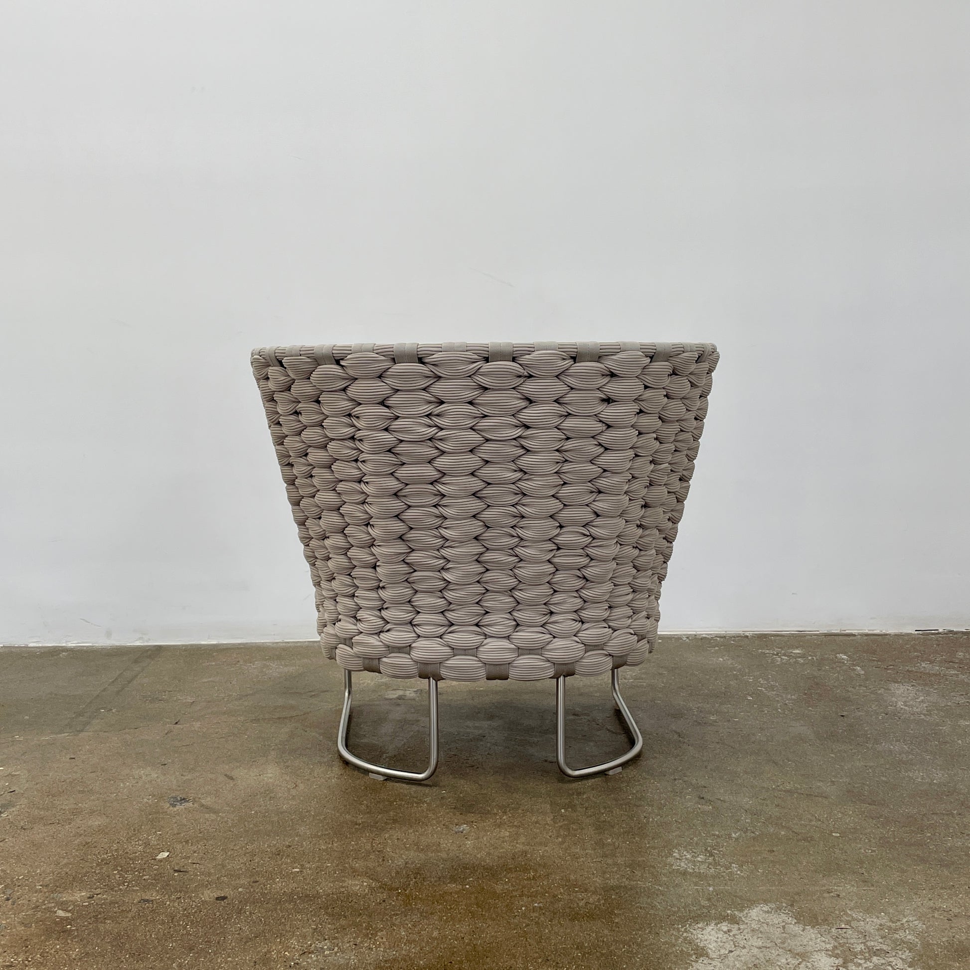 The Paola Lenti Ami Outdoor Armchair by Paola Lenti features a large weave frame, light gray cushions, and a metal base, all set against a plain white background.