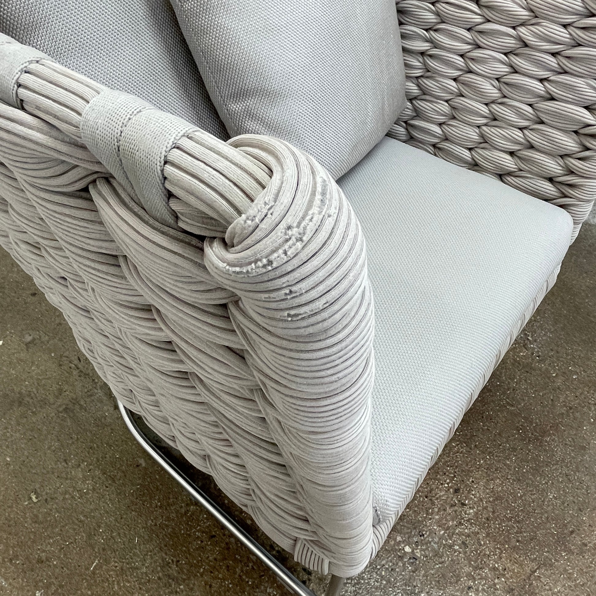 The Paola Lenti Ami Outdoor Armchair by Paola Lenti features a large weave frame, light gray cushions, and a metal base, all set against a plain white background.