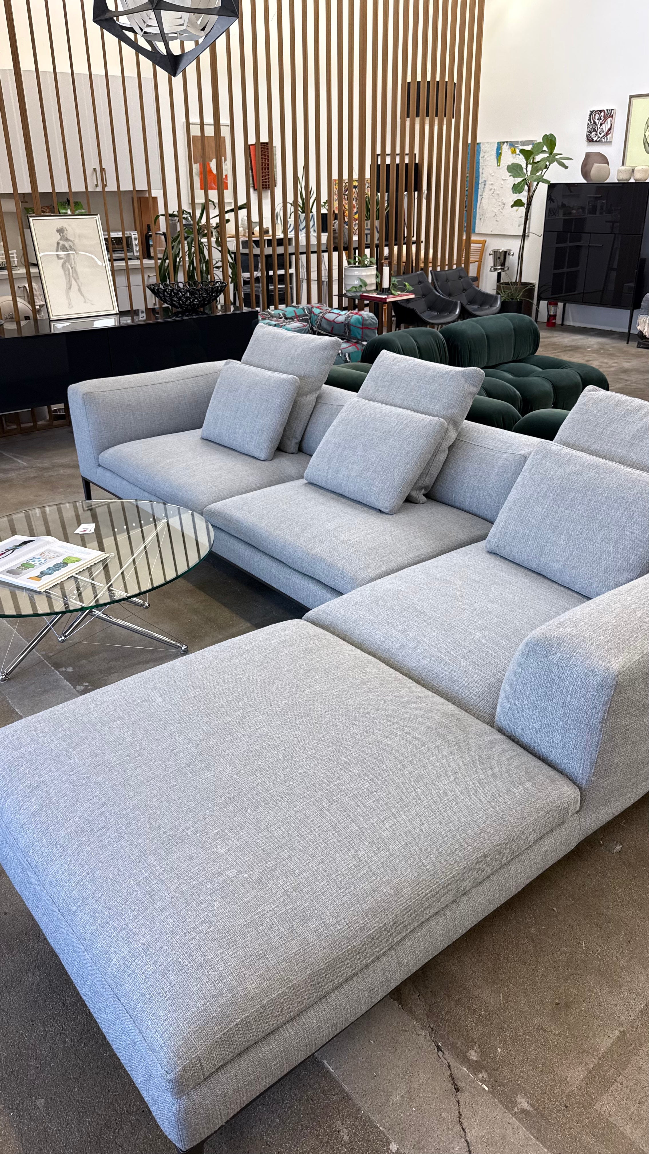 A modern living room features a large light gray B&B Italia Michel sectional from Modern Resale with cushions, a round glass coffee table, and other contemporary furniture on a concrete floor.
