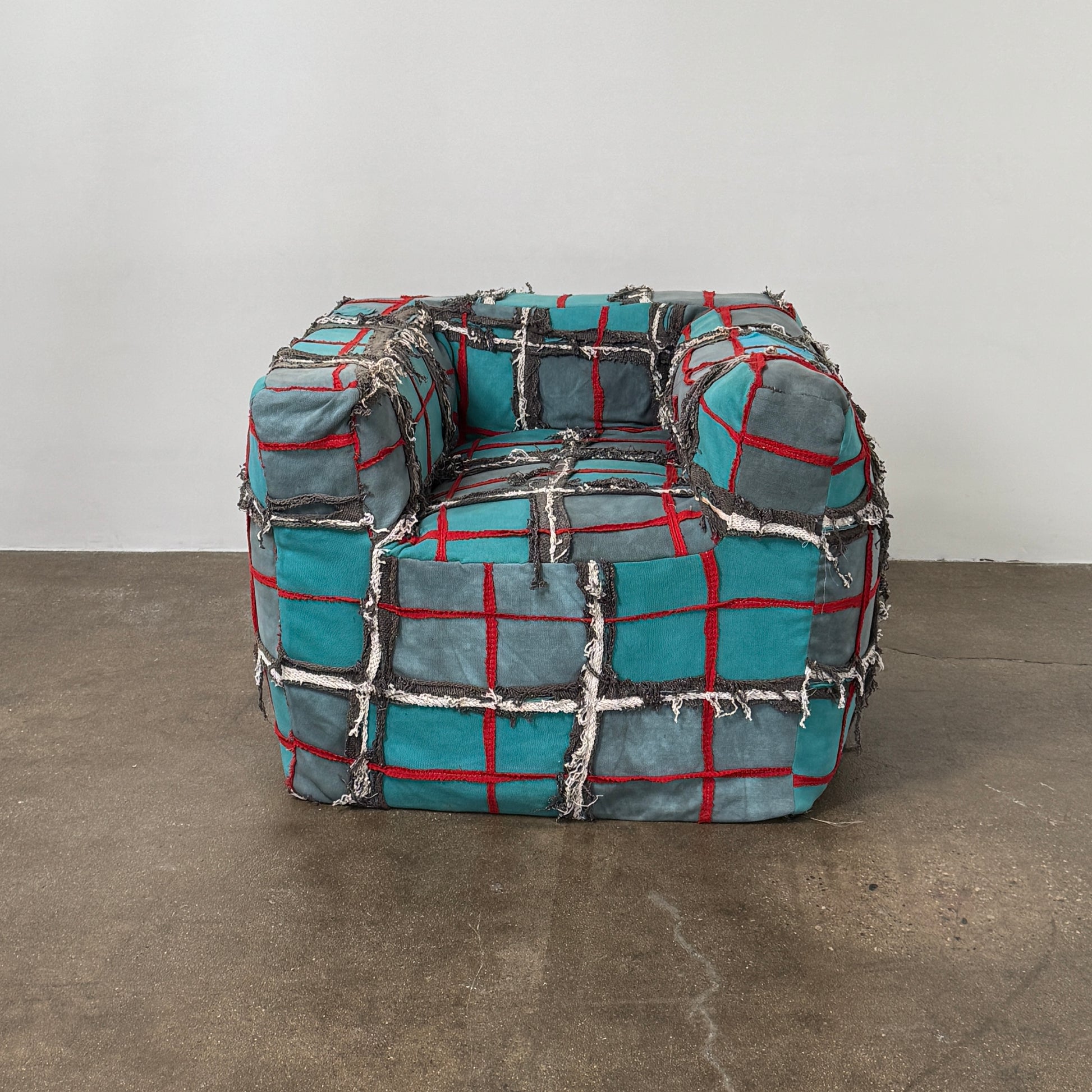 The Earth Landing Project Plaid Chair features boxy arms and patchwork teal, black, and red upholstery; frayed seams and irregular stitching create a rugged, textured look inspired by Earth Landing Project designs.