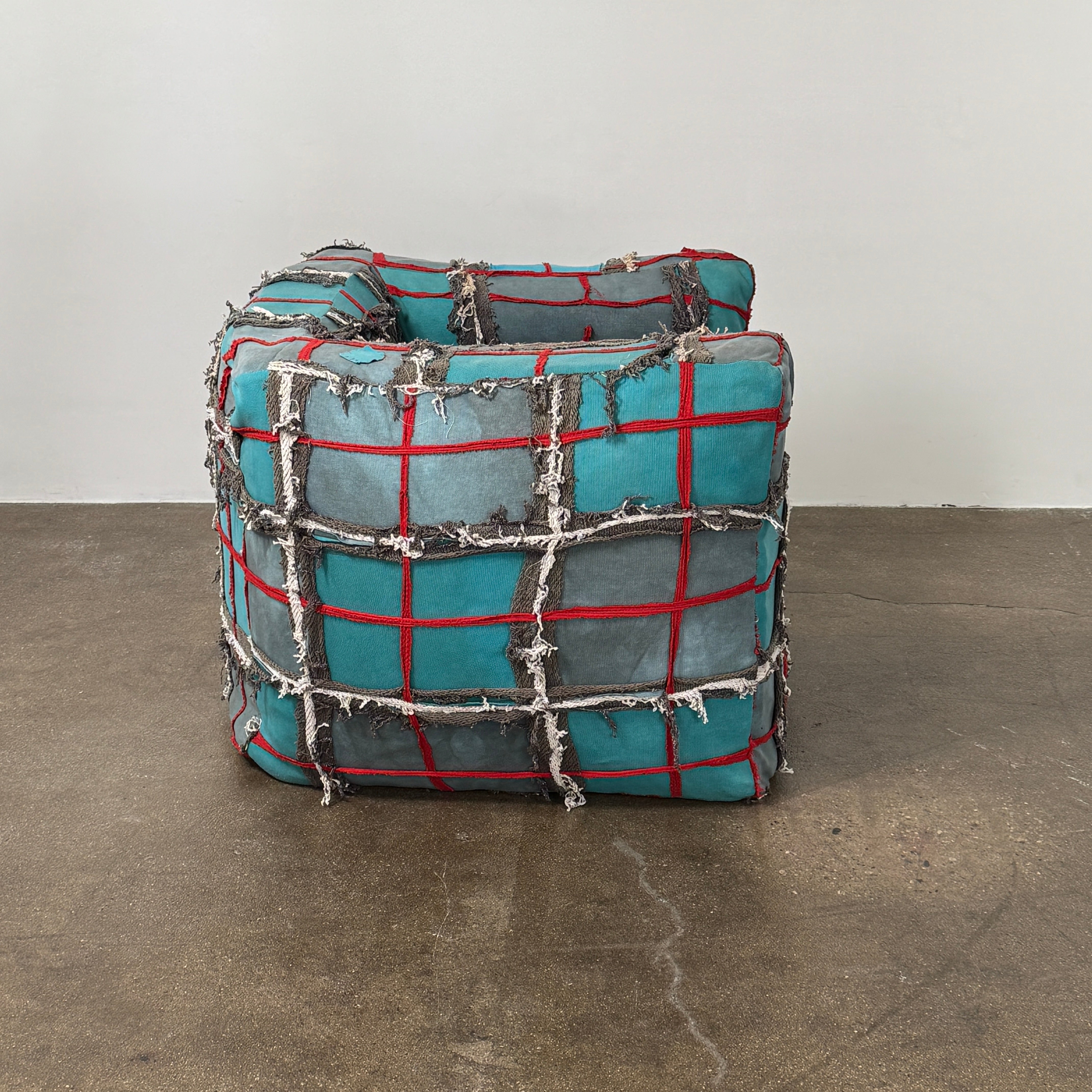 The Earth Landing Project Plaid Chair features boxy arms and patchwork teal, black, and red upholstery; frayed seams and irregular stitching create a rugged, textured look inspired by Earth Landing Project designs.