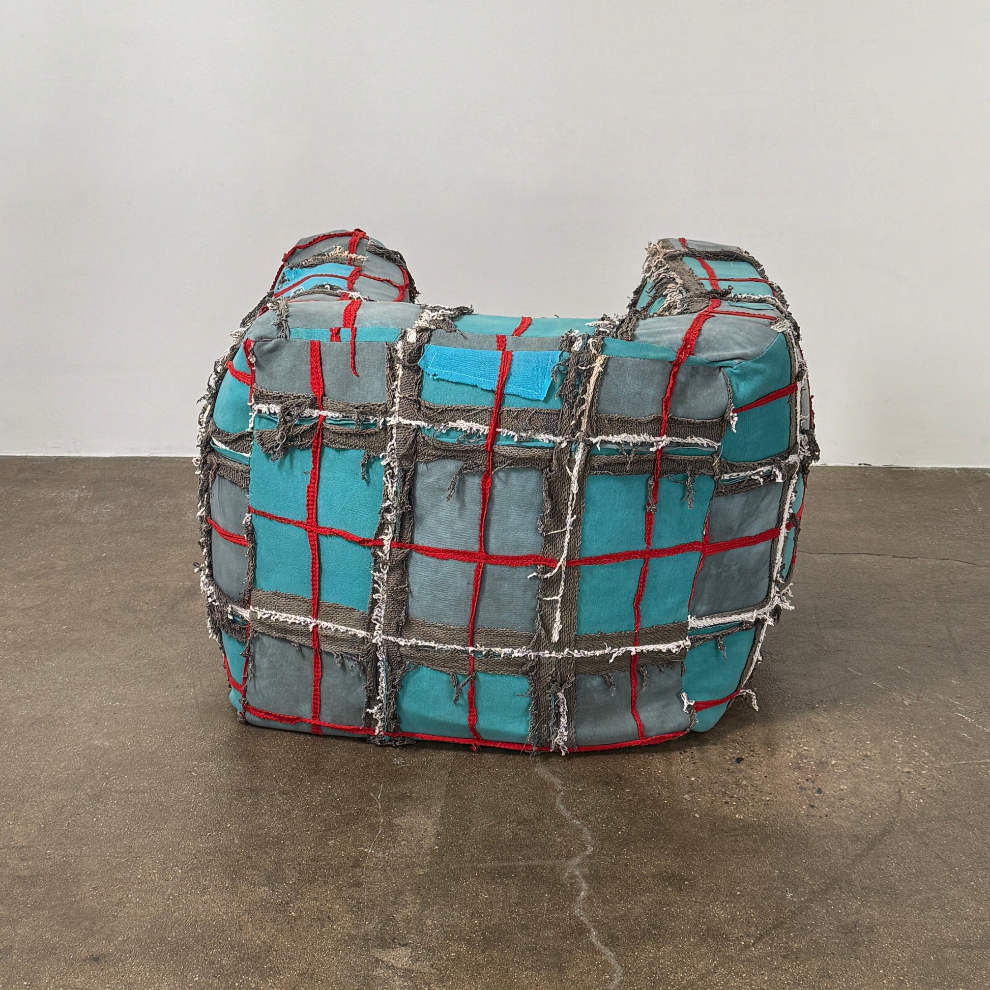 The Earth Landing Project Plaid Chair features boxy arms and patchwork teal, black, and red upholstery; frayed seams and irregular stitching create a rugged, textured look inspired by Earth Landing Project designs.