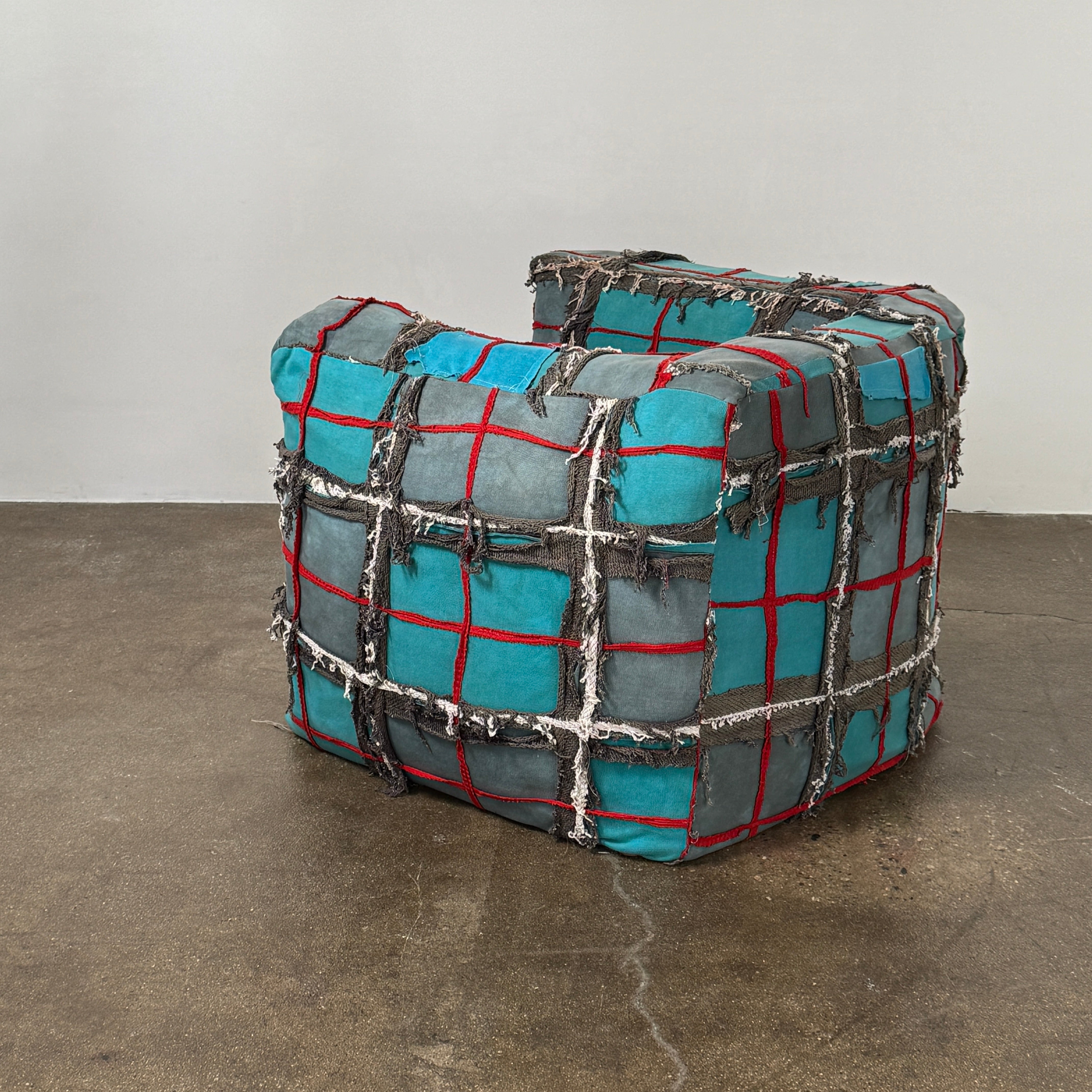 The Earth Landing Project Plaid Chair features boxy arms and patchwork teal, black, and red upholstery; frayed seams and irregular stitching create a rugged, textured look inspired by Earth Landing Project designs.
