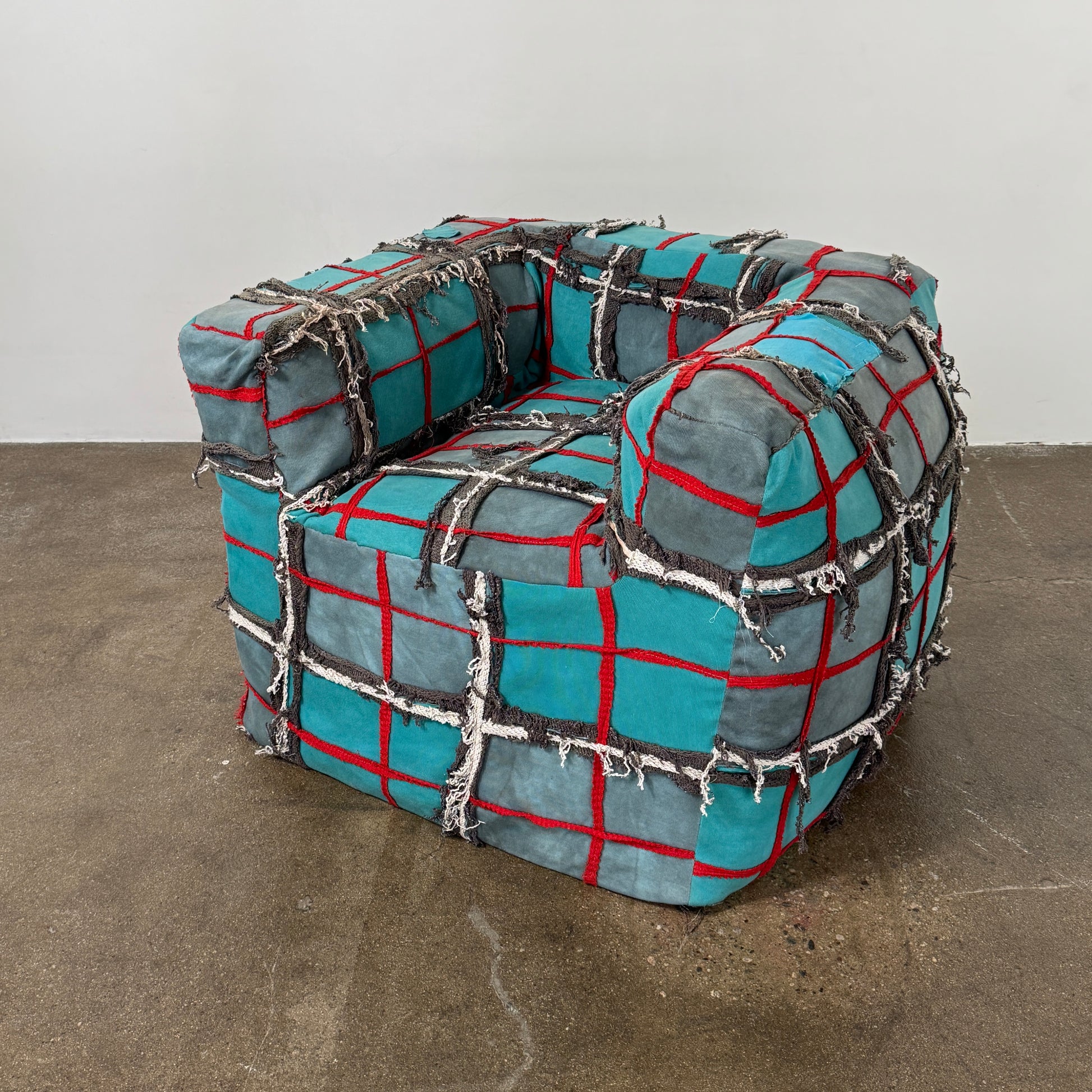 The Earth Landing Project Plaid Chair features boxy arms and patchwork teal, black, and red upholstery; frayed seams and irregular stitching create a rugged, textured look inspired by Earth Landing Project designs.
