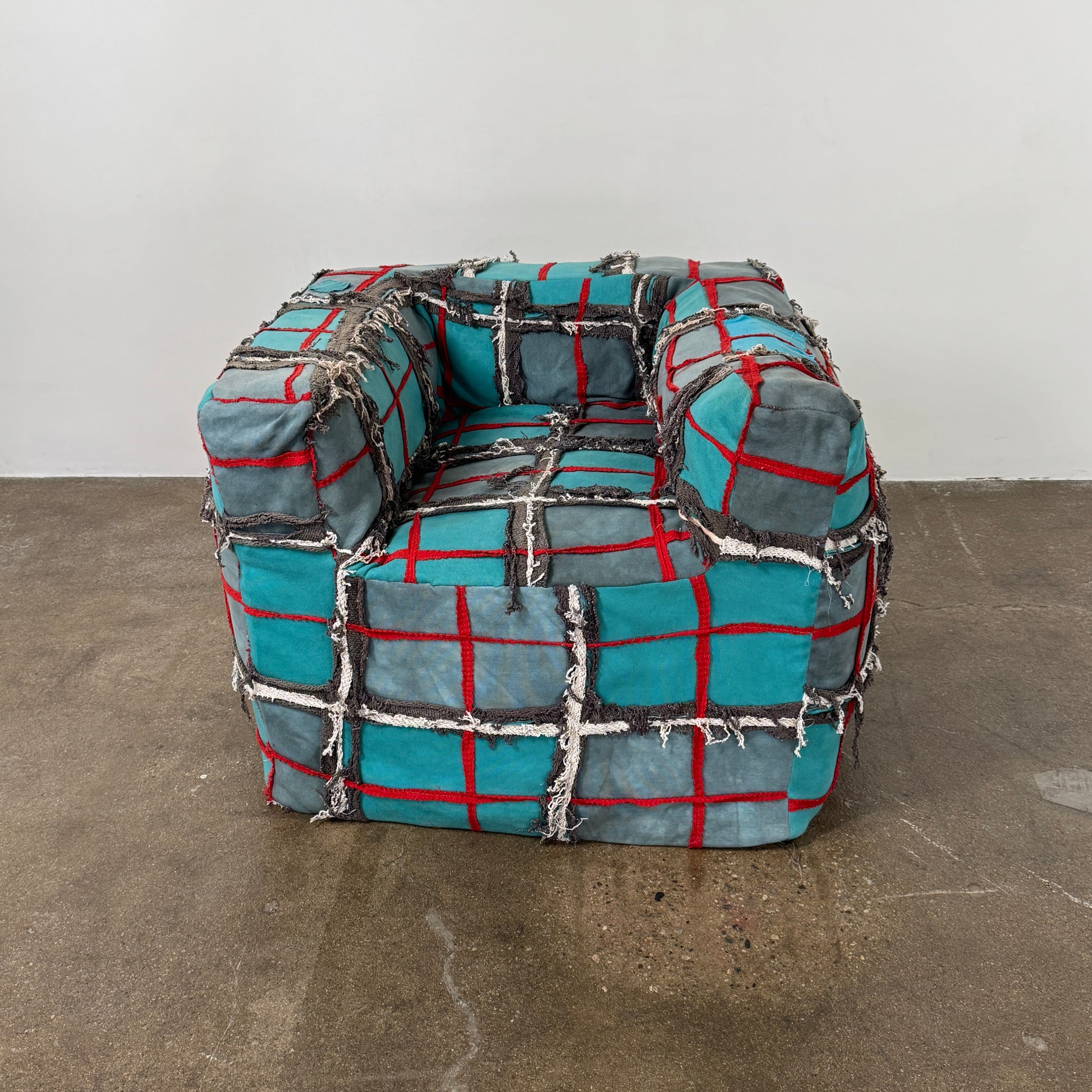 The Earth Landing Project Plaid Chair features boxy arms and patchwork teal, black, and red upholstery; frayed seams and irregular stitching create a rugged, textured look inspired by Earth Landing Project designs.