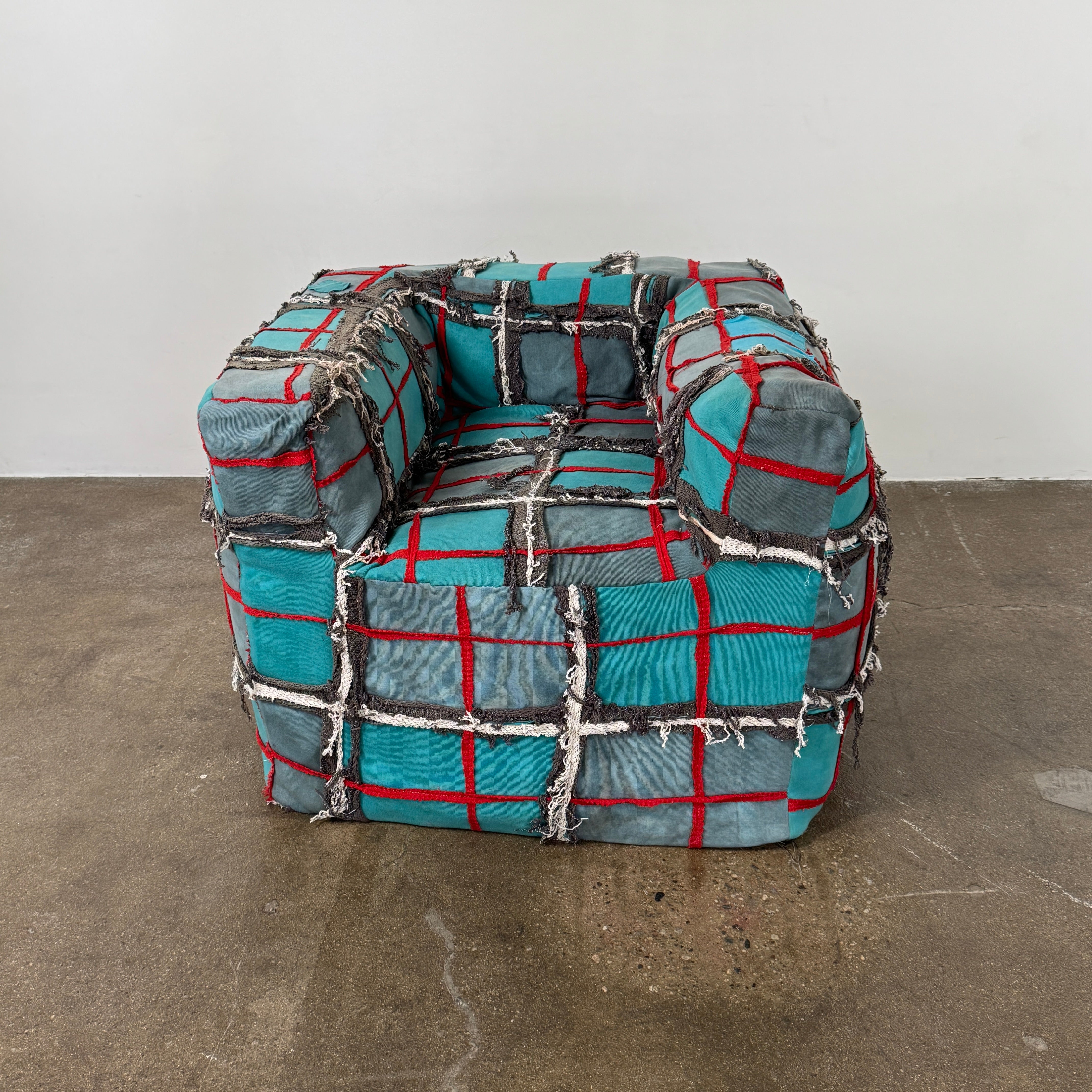 The Earth Landing Project Plaid Chair features boxy arms and patchwork teal, black, and red upholstery; frayed seams and irregular stitching create a rugged, textured look inspired by Earth Landing Project designs.