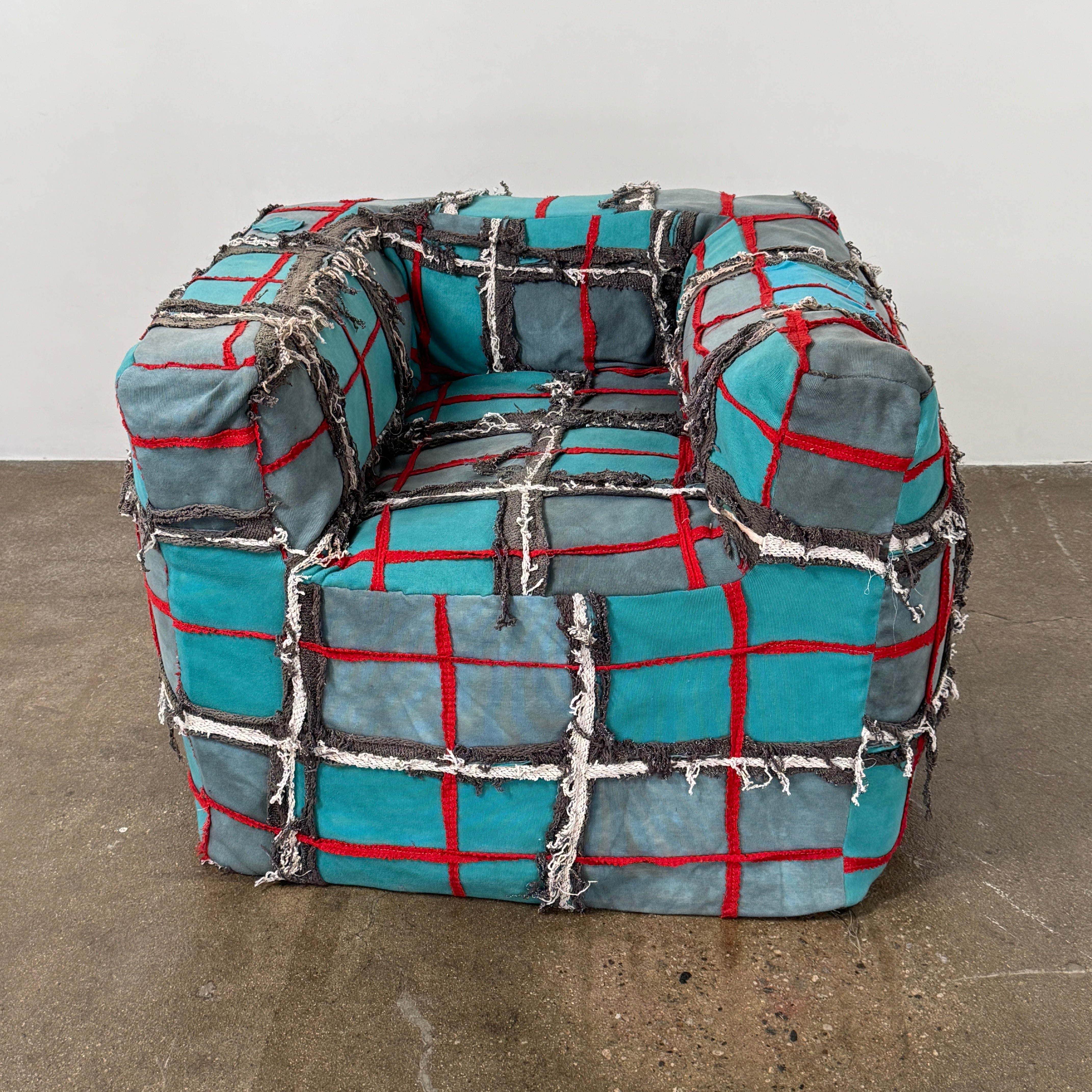 The Earth Landing Project Plaid Chair features boxy arms and patchwork teal, black, and red upholstery; frayed seams and irregular stitching create a rugged, textured look inspired by Earth Landing Project designs.