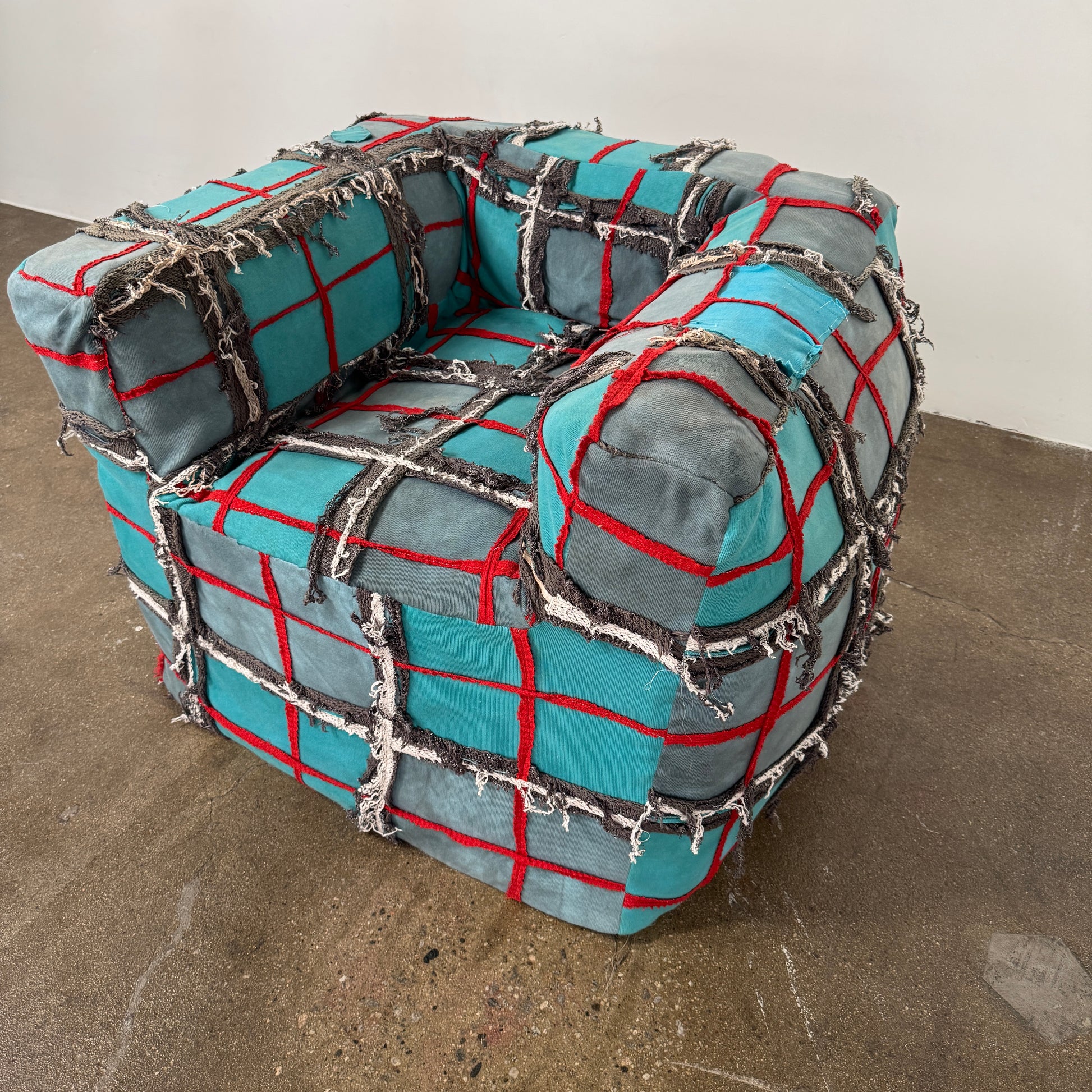 The Earth Landing Project Plaid Chair features boxy arms and patchwork teal, black, and red upholstery; frayed seams and irregular stitching create a rugged, textured look inspired by Earth Landing Project designs.