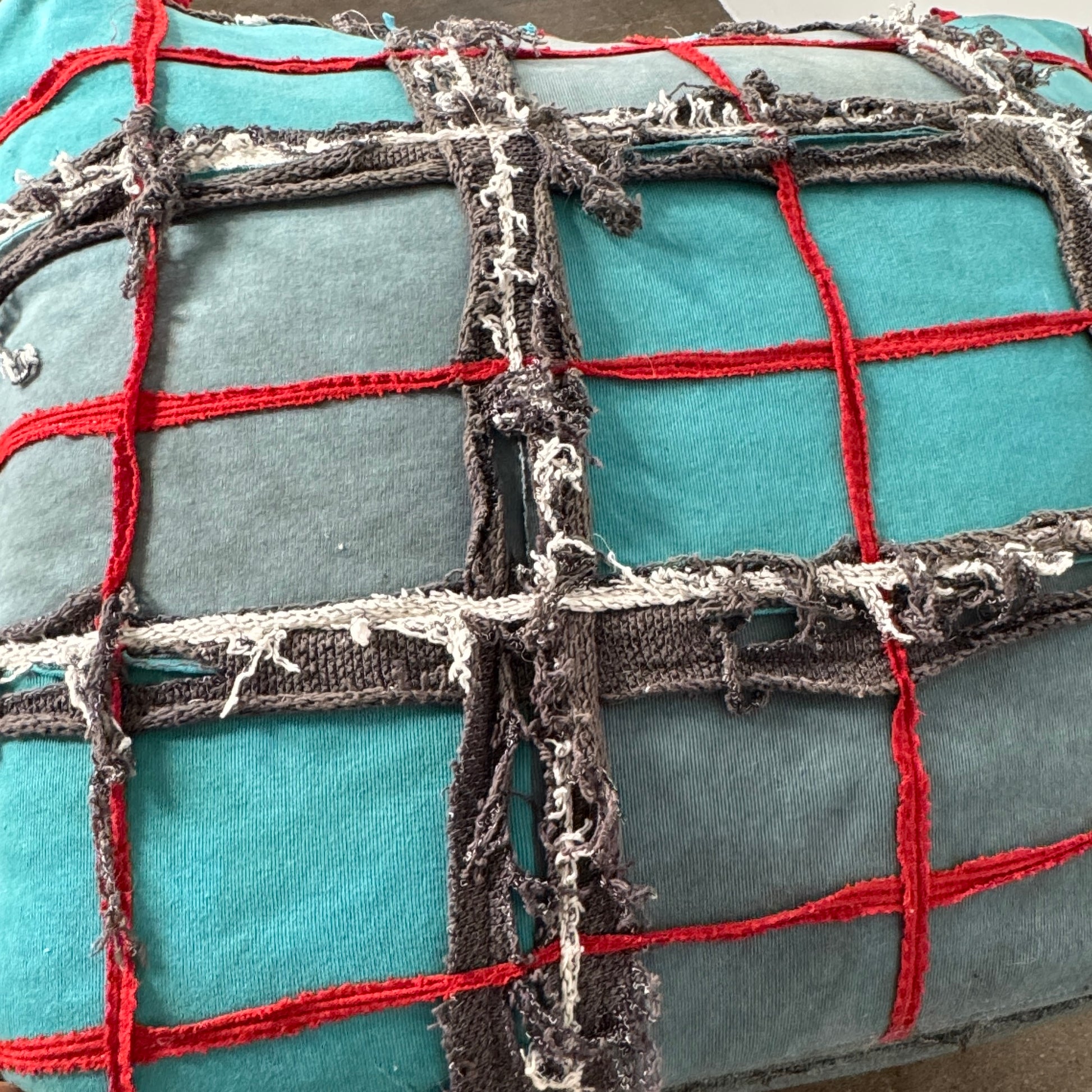 The Earth Landing Project Plaid Chair features boxy arms and patchwork teal, black, and red upholstery; frayed seams and irregular stitching create a rugged, textured look inspired by Earth Landing Project designs.