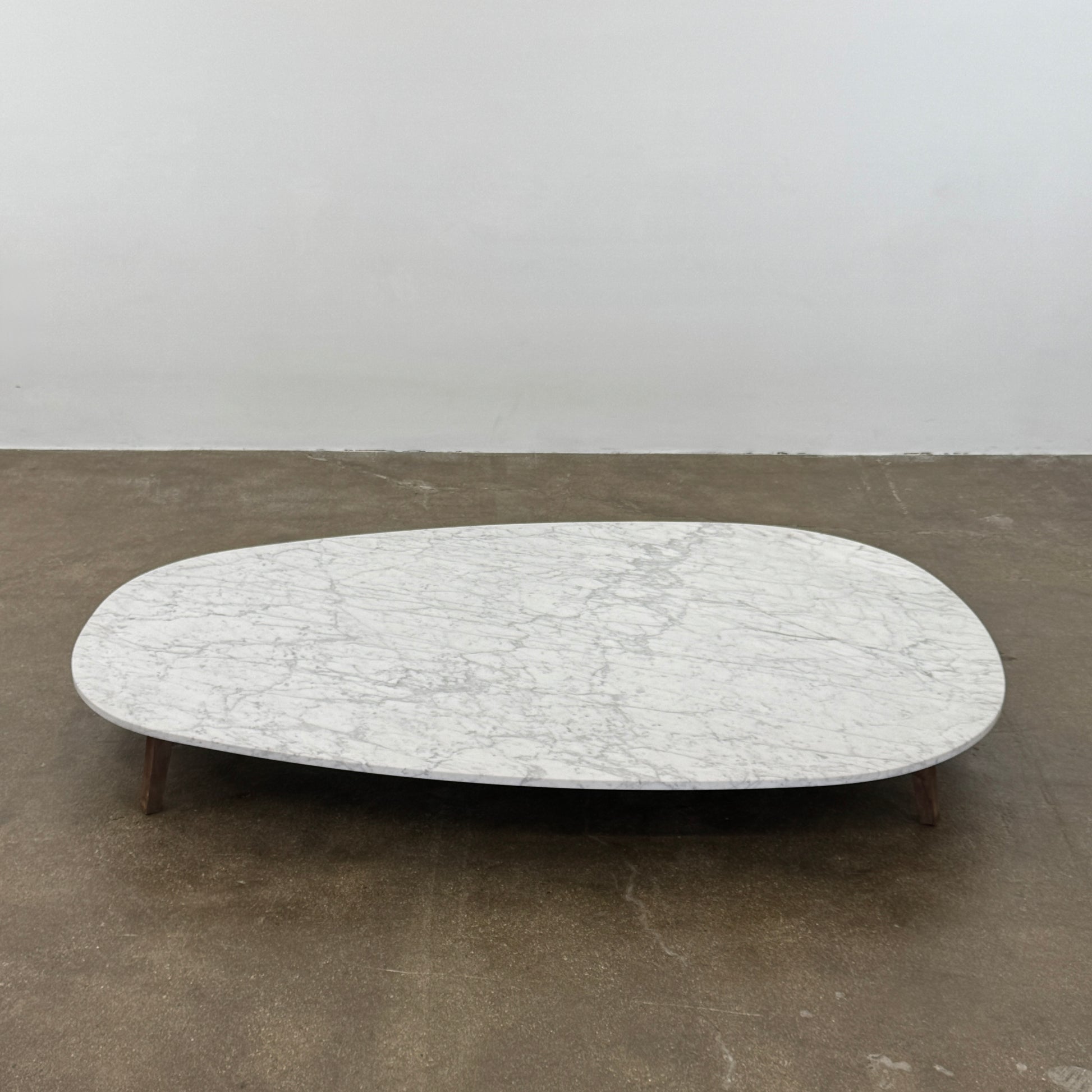 The Baxter Vietri Coffee Table by Baxter features a low, asymmetrical design with a white marble top and wooden legs, shown on a bare concrete floor against a plain white wall.