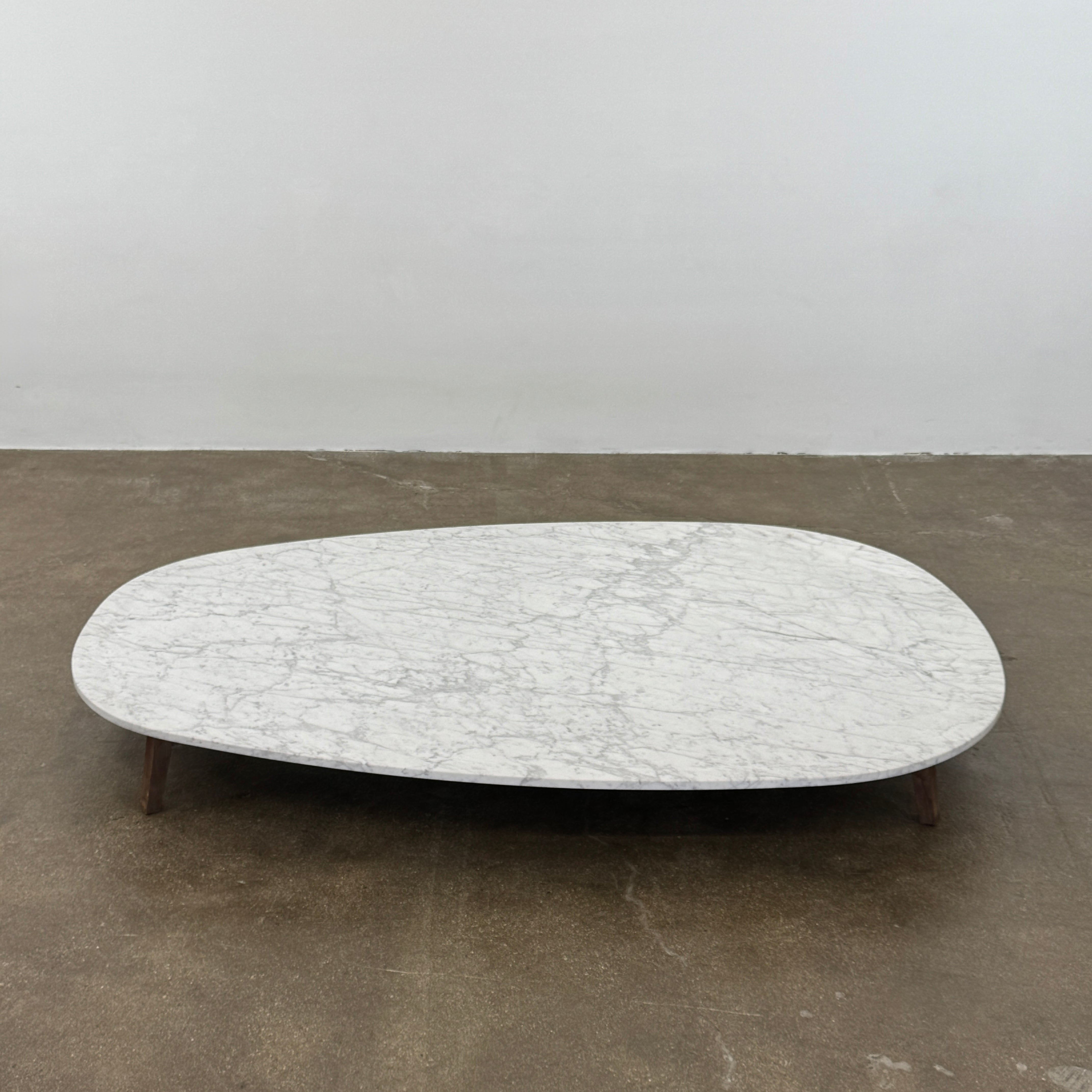 The Baxter Vietri Coffee Table by Baxter features a low, asymmetrical design with a white marble top and wooden legs, shown on a bare concrete floor against a plain white wall.