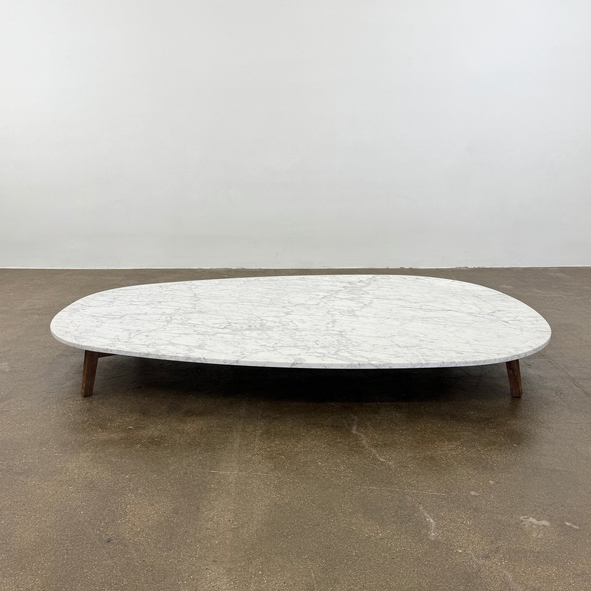 The Baxter Vietri Coffee Table by Baxter features a low, asymmetrical design with a white marble top and wooden legs, shown on a bare concrete floor against a plain white wall.