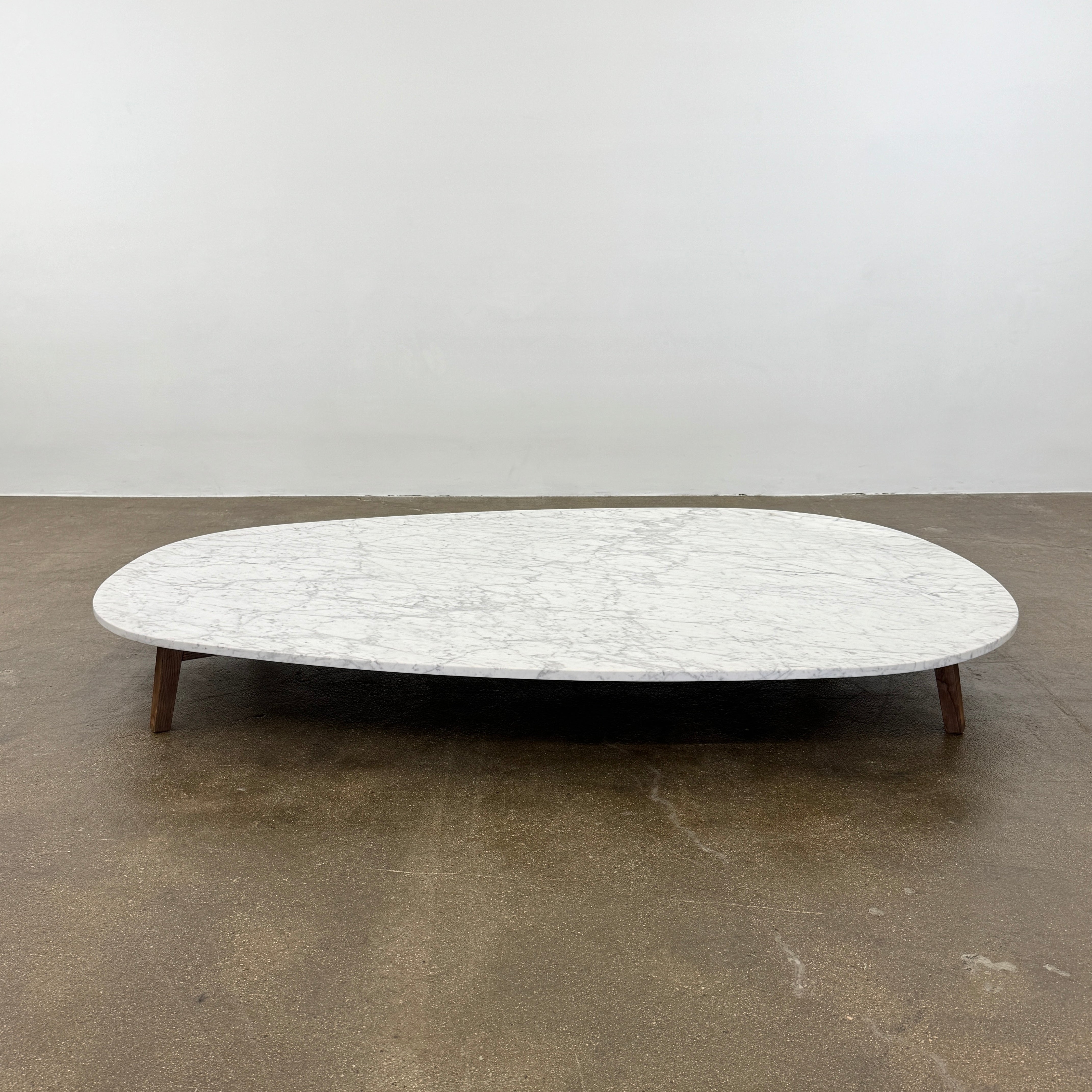The Baxter Vietri Coffee Table by Baxter features a low, asymmetrical design with a white marble top and wooden legs, shown on a bare concrete floor against a plain white wall.
