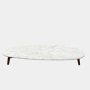 The Baxter Vietri Coffee Table by Baxter features a low, asymmetrical design with a white marble top and wooden legs, shown on a bare concrete floor against a plain white wall.
