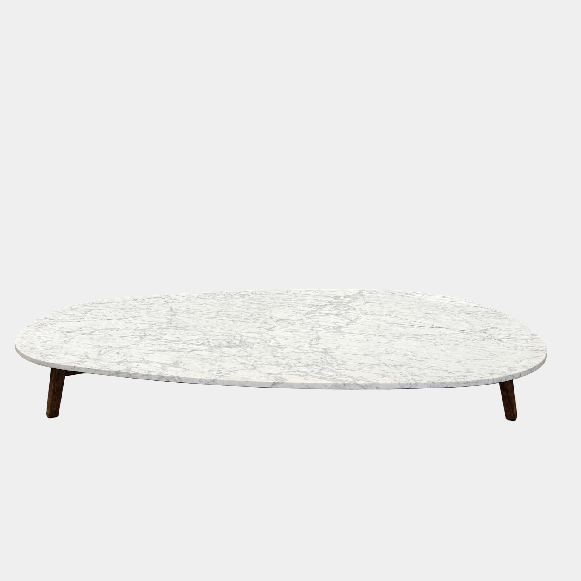 The Baxter Vietri Coffee Table by Baxter features a low, asymmetrical design with a white marble top and wooden legs, shown on a bare concrete floor against a plain white wall.