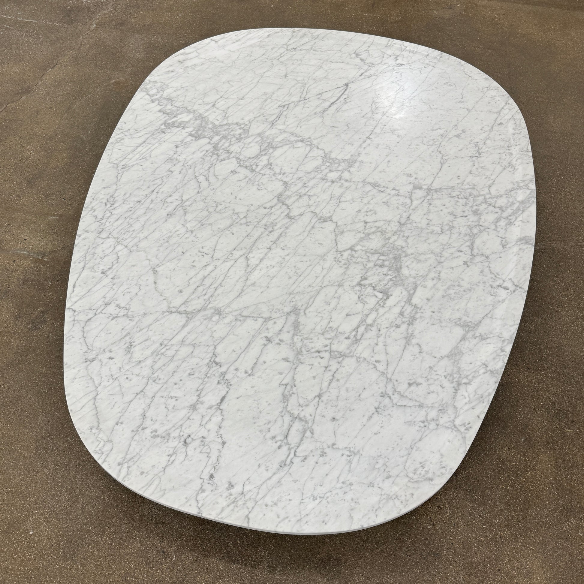 The Baxter Vietri Coffee Table by Baxter features a low, asymmetrical design with a white marble top and wooden legs, shown on a bare concrete floor against a plain white wall.