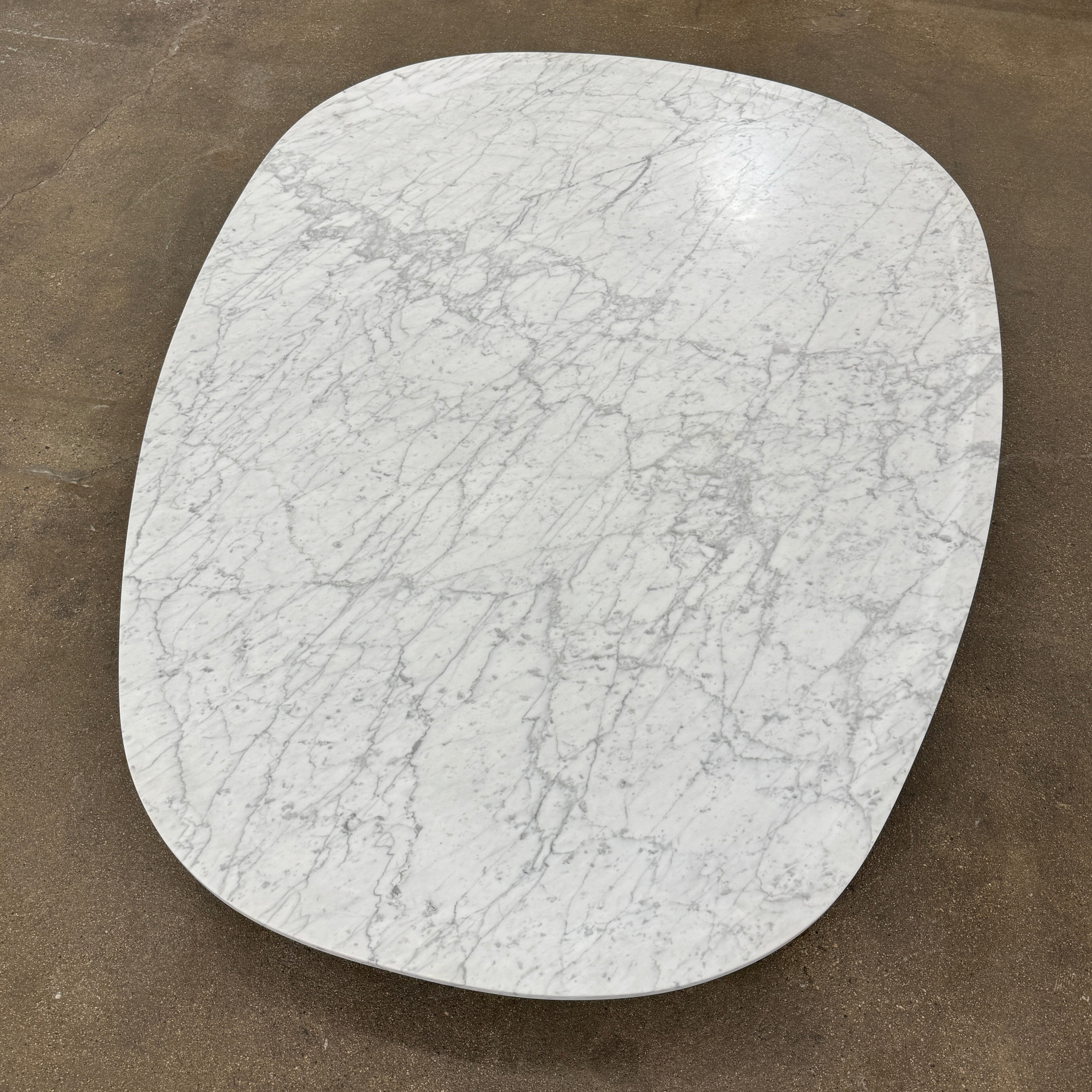 The Baxter Vietri Coffee Table by Baxter features a low, asymmetrical design with a white marble top and wooden legs, shown on a bare concrete floor against a plain white wall.