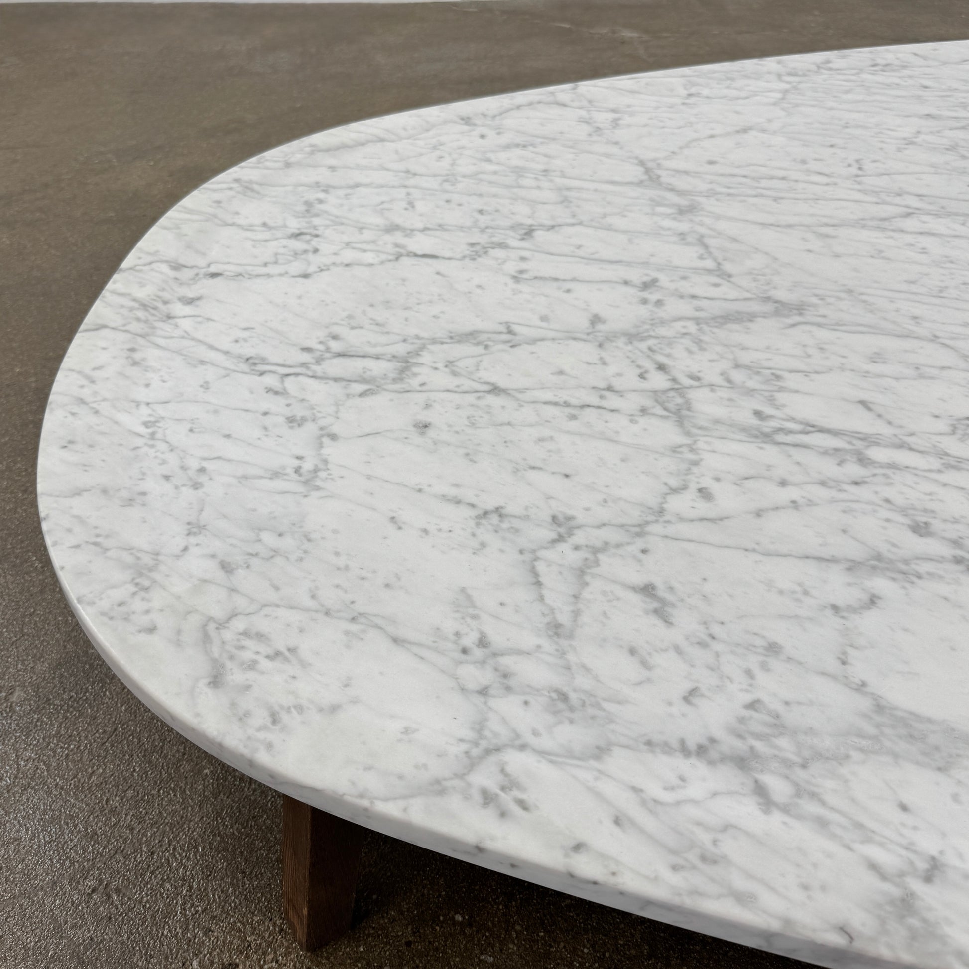 The Baxter Vietri Coffee Table by Baxter features a low, asymmetrical design with a white marble top and wooden legs, shown on a bare concrete floor against a plain white wall.