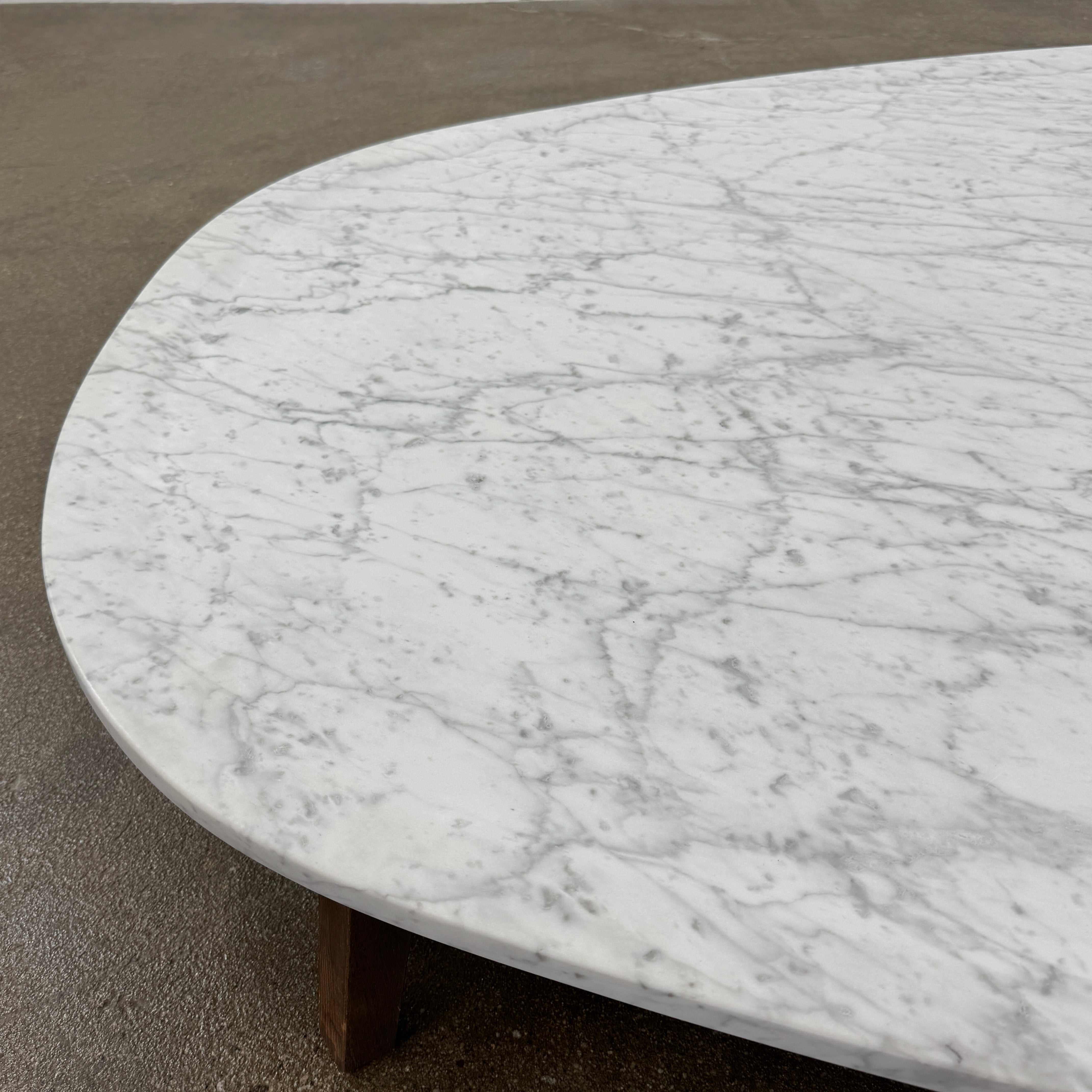 The Baxter Vietri Coffee Table by Baxter features a low, asymmetrical design with a white marble top and wooden legs, shown on a bare concrete floor against a plain white wall.