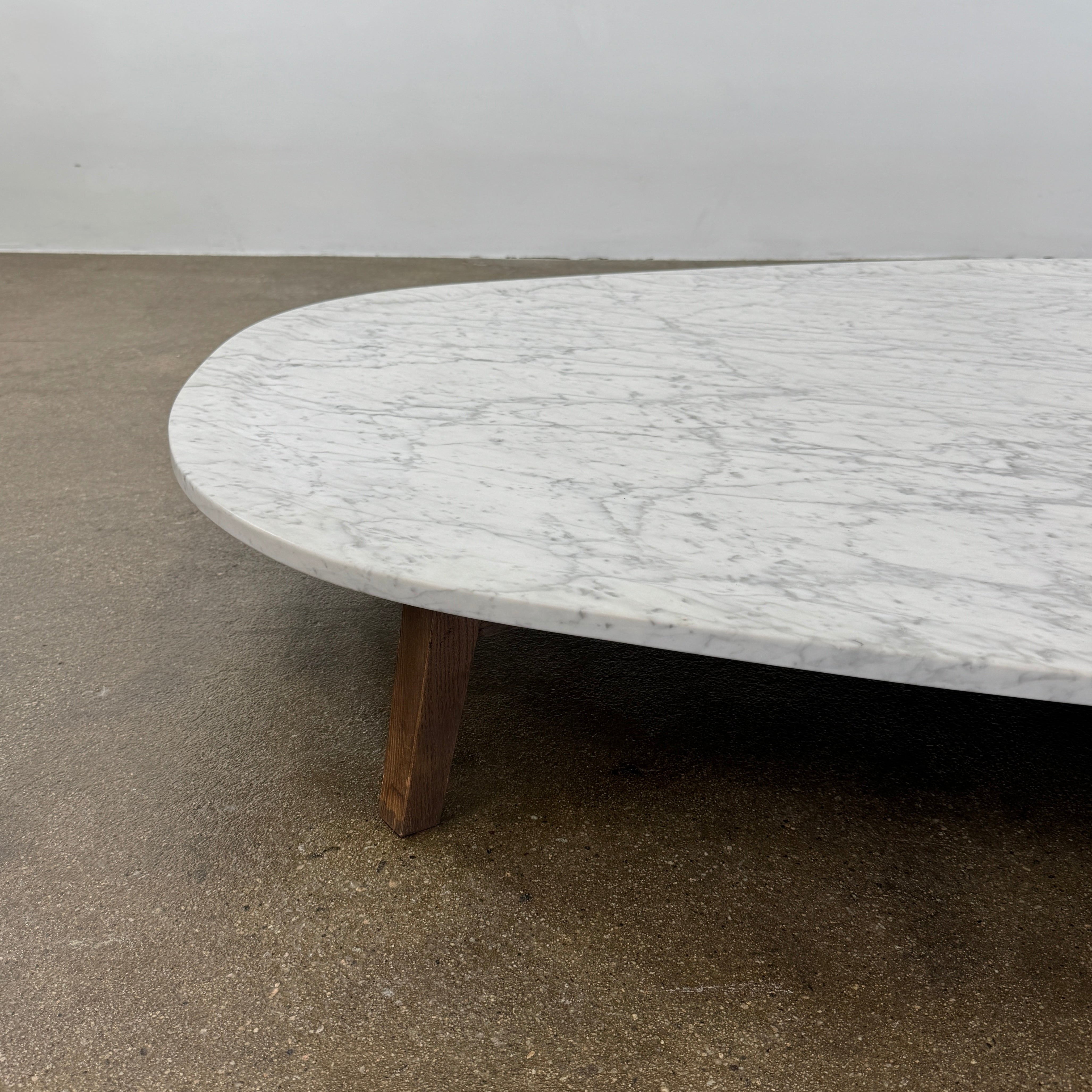 The Baxter Vietri Coffee Table by Baxter features a low, asymmetrical design with a white marble top and wooden legs, shown on a bare concrete floor against a plain white wall.