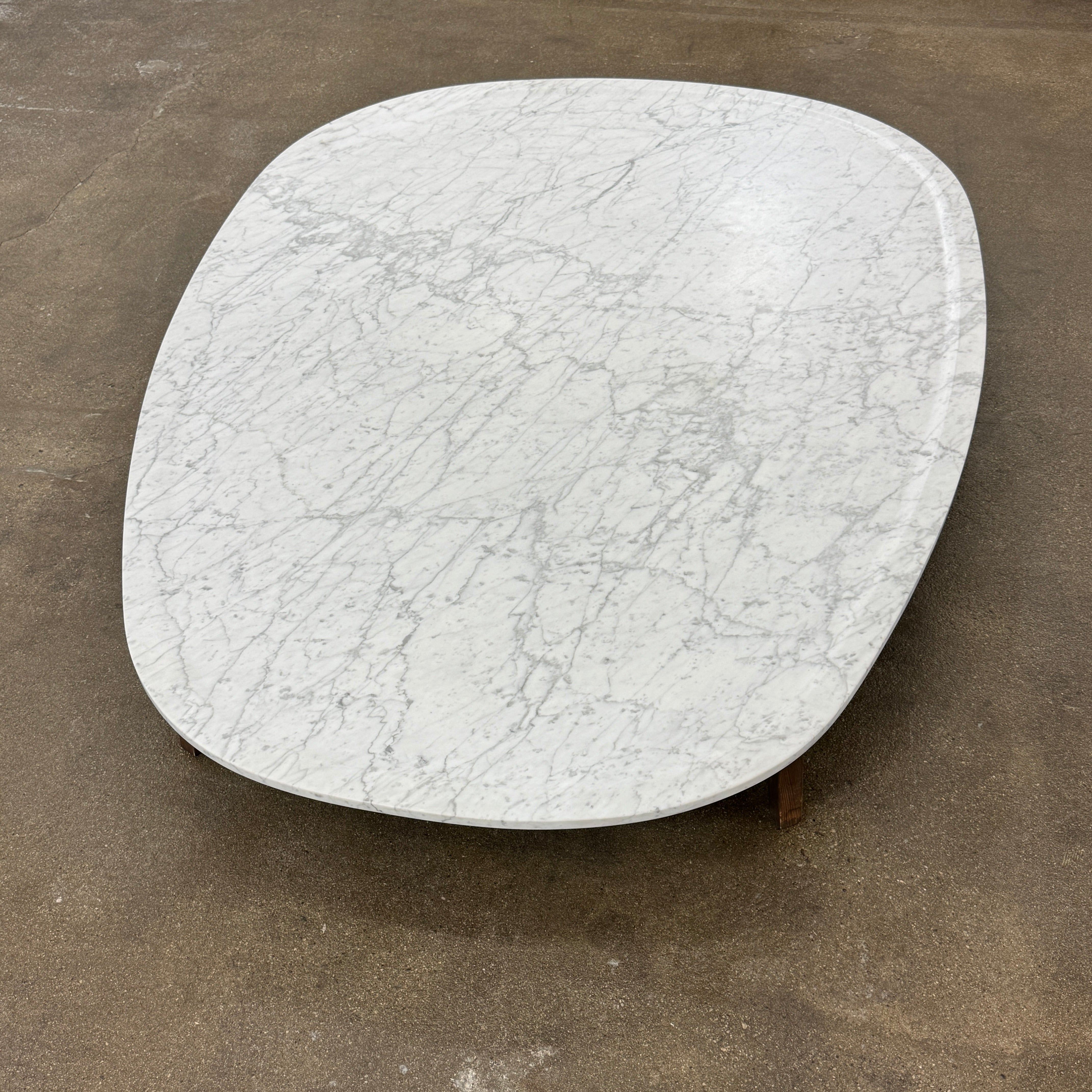 The Baxter Vietri Coffee Table by Baxter features a low, asymmetrical design with a white marble top and wooden legs, shown on a bare concrete floor against a plain white wall.