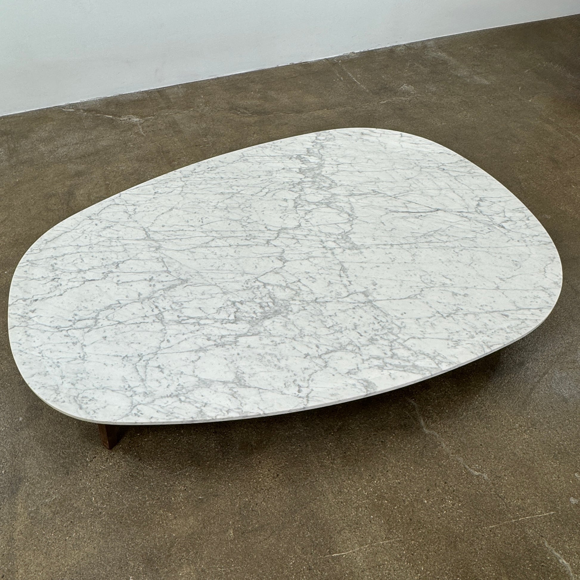 The Baxter Vietri Coffee Table by Baxter features a low, asymmetrical design with a white marble top and wooden legs, shown on a bare concrete floor against a plain white wall.