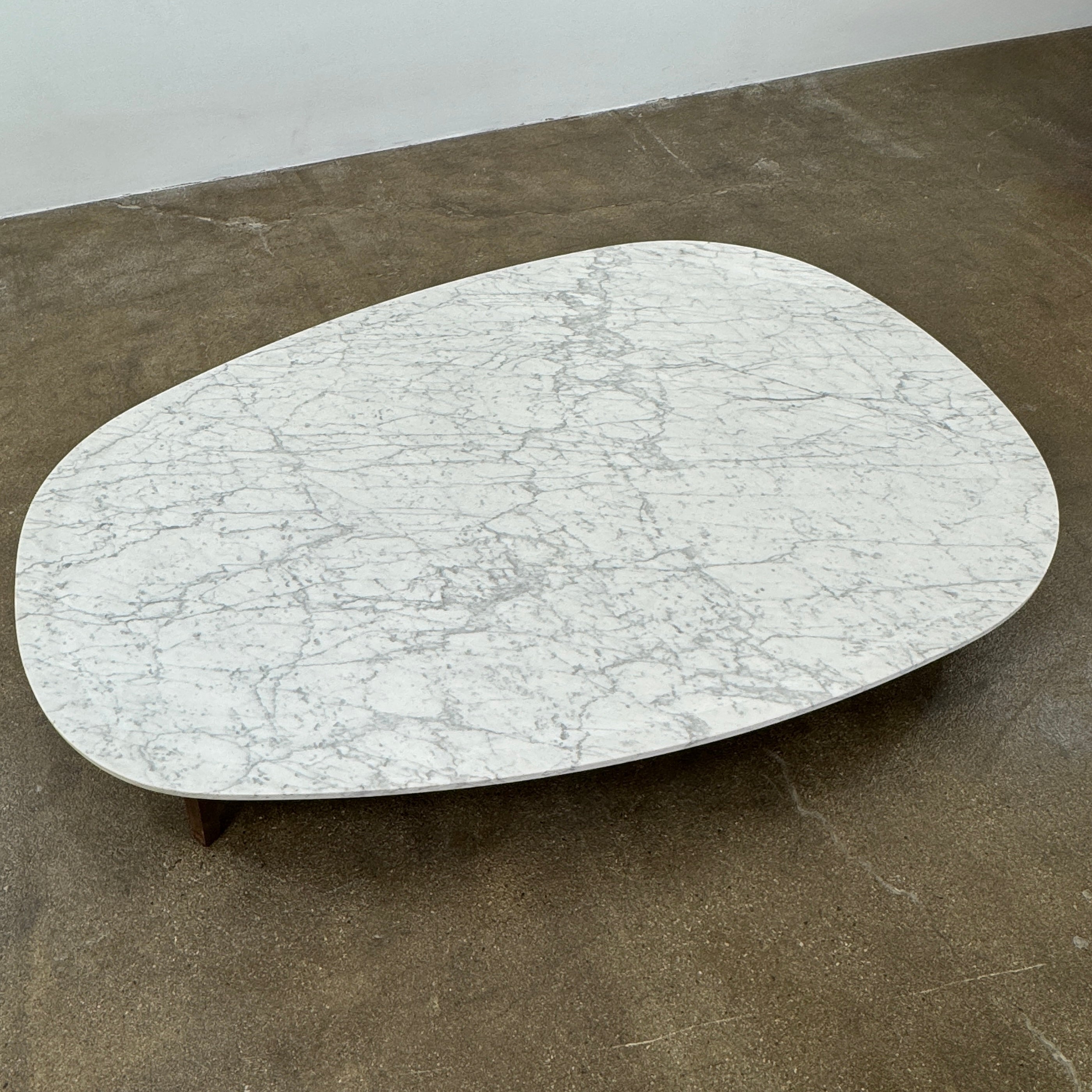 The Baxter Vietri Coffee Table by Baxter features a low, asymmetrical design with a white marble top and wooden legs, shown on a bare concrete floor against a plain white wall.