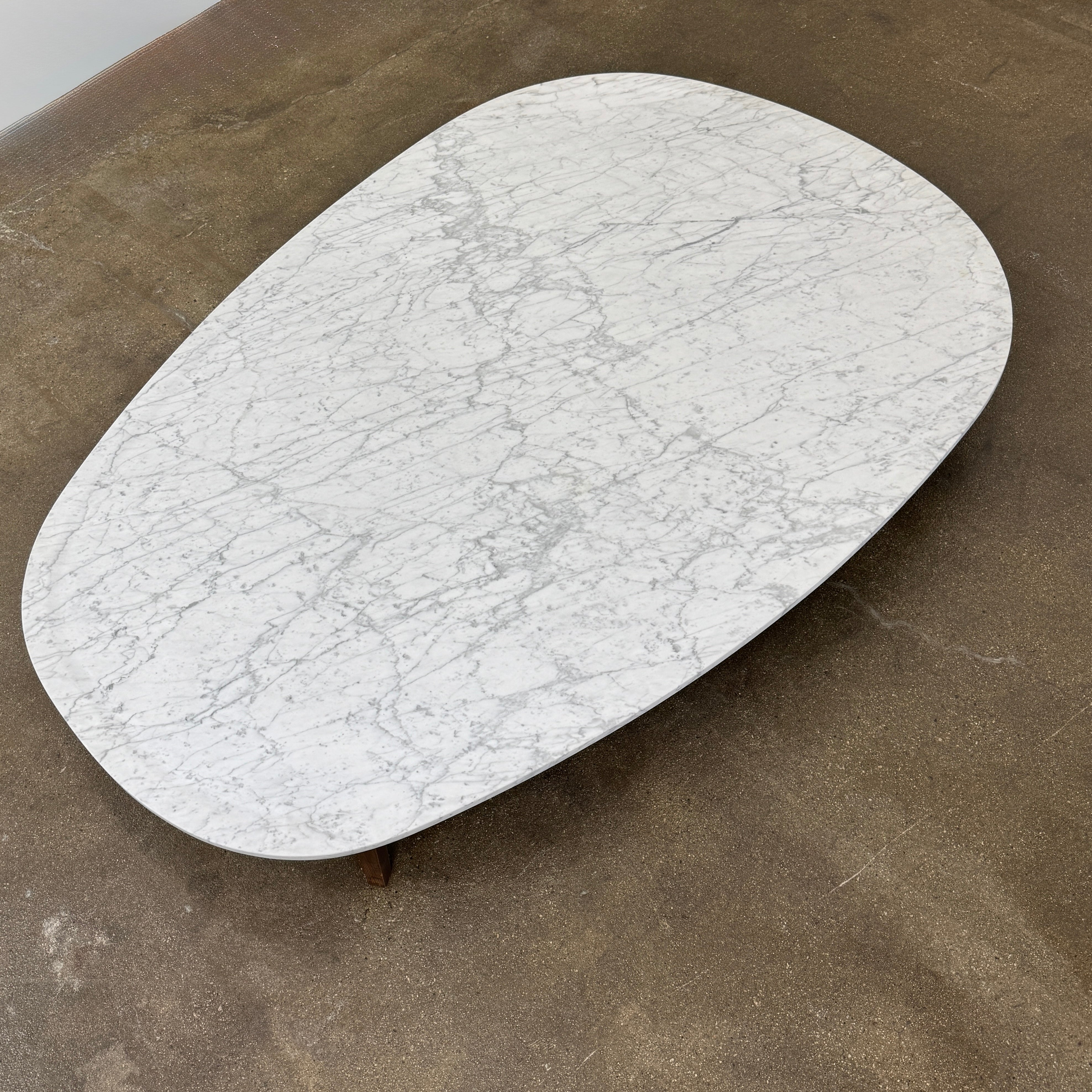 The Baxter Vietri Coffee Table by Baxter features a low, asymmetrical design with a white marble top and wooden legs, shown on a bare concrete floor against a plain white wall.